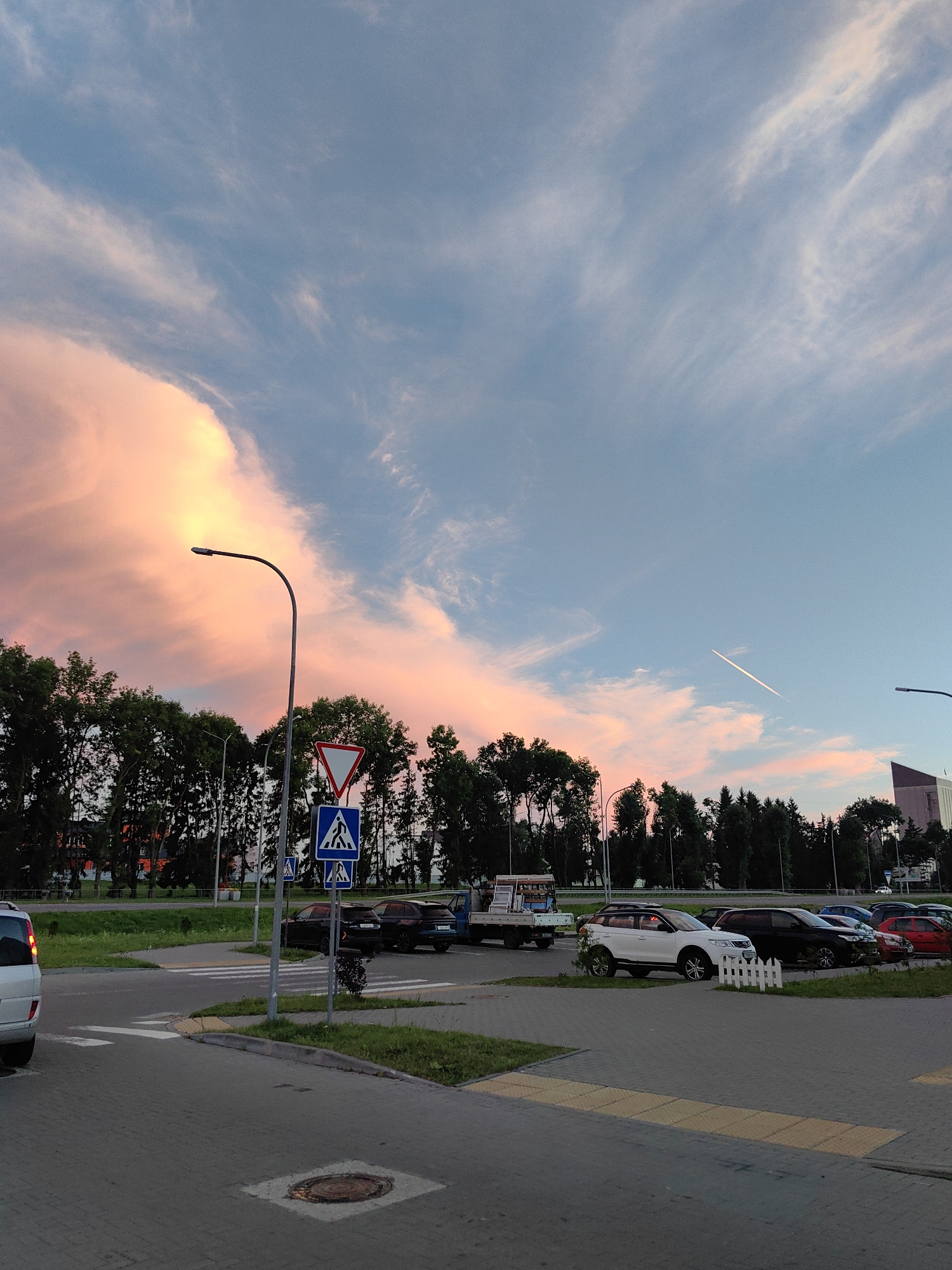 The image depicts a street scene at dusk in Dzyarzhynsk, Belarus, characterized by a vibrant sky painted with hues of pink, orange, and blue, with wispy clouds and a faint contrail from an airplane. In the foreground, a paved area with pedestrian crossings and road markings is visible, along with a manhole cover. Several vehicles are parked in a lot adjacent to the road, including a white SUV, a black SUV, and a red car. A small white picket fence separates the parking area from a grassy strip. A truck with a load is also present. Road signs indicating pedestrian crossings and a yield sign are positioned near a street lamp. A dense line of trees forms a backdrop, with buildings visible in the distance. The scene suggests a typical urban environment during twilight.