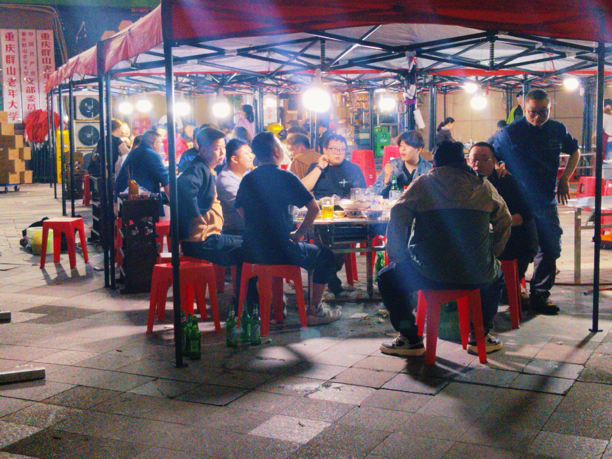 This image shows a group of people dining outdoors under a series of canopies supported by a black metal frame structure, likely at night. The scene is brightly lit by numerous hanging light bulbs.
Several individuals are seated at a table laden with food and drinks, while others are standing. Many of them are male. They appear to be engaged in conversation and socializing. Some are eating or drinking.
The setting appears to be an outdoor food stall or gathering space in the city of Yuzhong District, China. There are red plastic stools scattered around the area, some occupied and some empty. Green glass beer bottles are visible on the ground, suggesting consumption of alcohol. In the background, there are signs in Chinese characters, including text that translates to "Chongqing Qunshan Elderly University". There are also what appears to be air conditioning units on the left and storage boxes.
The ground is paved with grey tiles. Shadows of the canopies and support structures are cast onto the pavement. The overall atmosphere suggests a casual, social gathering in a public space.
