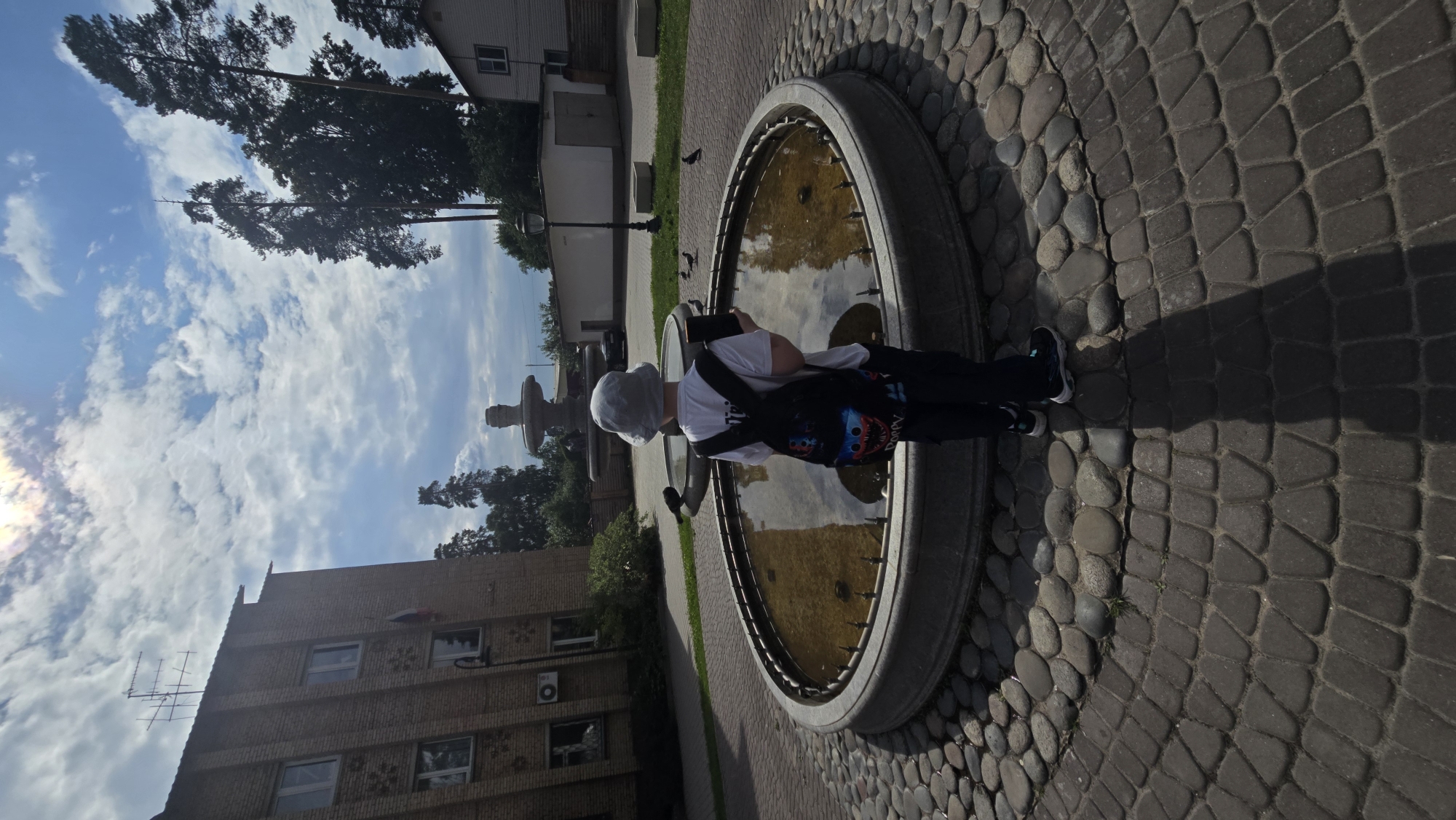 A person wearing a light-colored hat, white t-shirt, and dark pants stands by a circular fountain filled with water. The fountain is surrounded by paving stones, a mix of large, rounded ones and smaller, rectangular ones. The person is holding a dark object, possibly a phone or tablet, and has a backpack with a colorful design over their shoulder.
The scene appears to be outdoors in a park or public square. In the background, there's a building made of brick with several windows and an air conditioning unit visible. A fountain with multiple tiers is also present in the background, further away from the main subject. Tall trees with dark green foliage frame the scene, and a bright blue sky with scattered white clouds suggests it is daytime, likely sunny. The sun is casting a noticeable shadow on the paving stones.
The overall atmosphere seems peaceful, with the person engaged in their own activity. The presence of birds near the fountain suggests a natural environment. The location is identified as Il’inskiy, Russia. There is no visible text in the image.