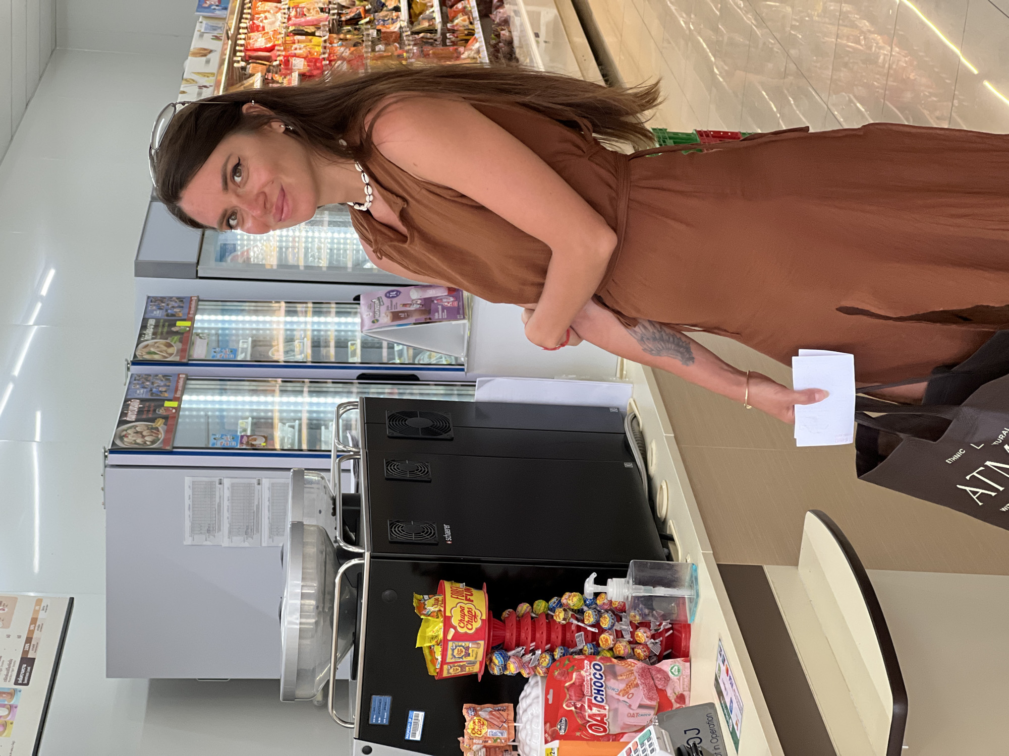 This media file features a woman in a brown dress standing in what appears to be a convenience store or a small supermarket. The woman is positioned in the center of the frame, looking towards the camera with a slight smile. She is wearing a seashell necklace and has sunglasses perched on her head. Her right arm is visible, and there is a tattoo on her left forearm. She is holding a white piece of paper in her right hand.
The background reveals rows of refrigerated display cases filled with food items, suggesting a retail environment. To the right of the woman, a black machine with fans is visible, and in front of it is a display of Chupa Chups lollipops and other snack items, including "OAT CHOCO" and "HIGH PROTEIN" products. A dark shopping bag with the text "ETHNIC L AT" is also visible near the right edge of the frame.
The lighting appears to be artificial, consistent with an indoor setting. The overall atmosphere suggests a casual shopping trip. The visible text includes product names and brand logos. The location context provided indicates the setting is Ko Pha Ngan, Thailand. The image is oriented vertically.