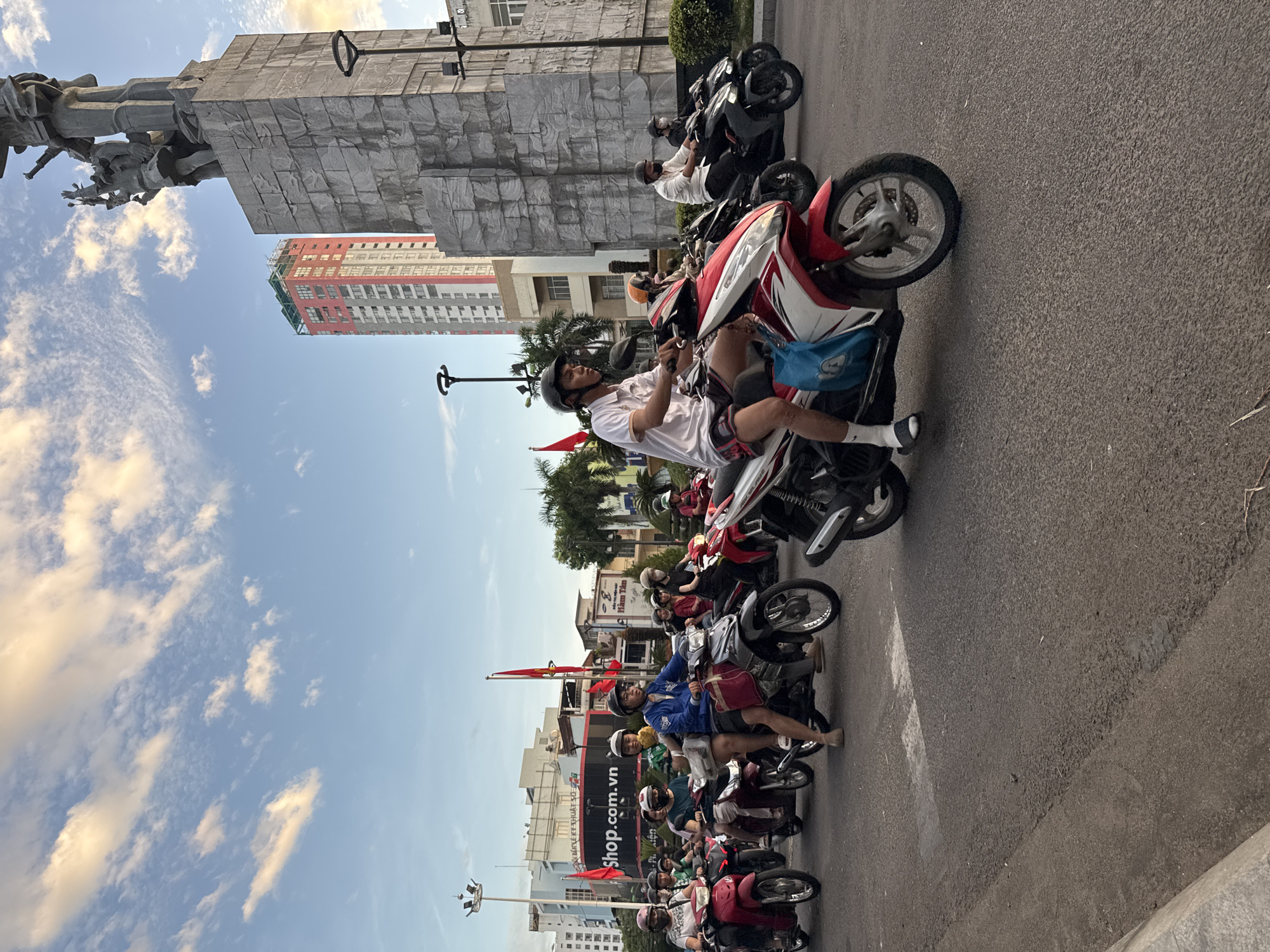 A wide shot captures a street scene in Nha Trang, Vietnam, with a group of people riding motorcycles under a cloudy sky with hints of golden sunlight. In the background, a large, stone monument with sculpted figures stands prominently, adjacent to a tall, red and white building. The street is paved, and there are various buildings and palm trees lining the sides. Red flags are visible in the distance, and a sign reading "Shop.com.vn" is partially obscured by the motorcycles. The motorcycles are diverse in color, including red, white, black, and silver. Most riders are wearing helmets. The overall impression is one of everyday urban life and movement.