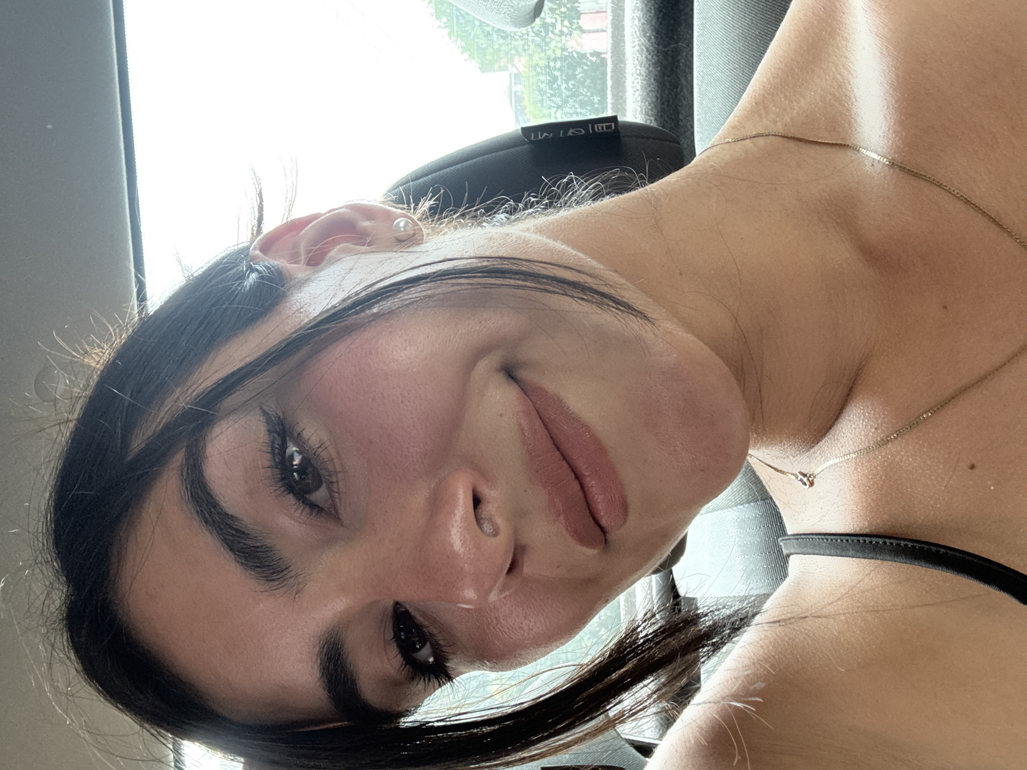 A close-up, rotated image depicts a young woman with fair skin, dark hair, and brown eyes. She is wearing a pearl earring in her left ear and a gold chain necklace around her neck. The woman has full lips and noticeable eyelashes. A black strap is visible on her right shoulder. The background is out of focus, but appears to show a car interior with a window, suggesting the location is likely inside a vehicle. There is no specific information to determine the location in the city of Puebla, Mexico.