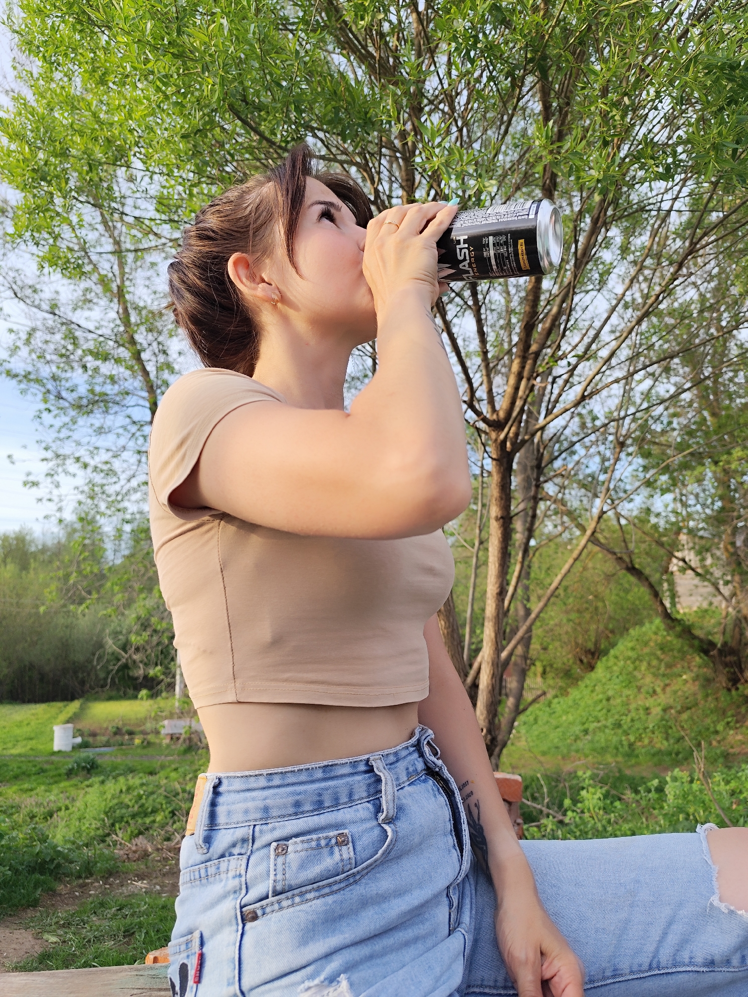 A young woman drinks from a can outdoors. She has brown hair styled in an updo with loose strands framing her face, and is wearing a cropped, light brown t-shirt and light blue, distressed denim shorts. She is seated, possibly on a bench, with her knees up and one arm bent as she drinks. The can she's holding is black with silver accents, and has the words "FLASH ENERGY" visible. The background consists of a green, grassy area with trees, suggesting a park-like setting. The location may be Фоки, Russia, based on the query information. The lighting appears to be natural daylight. A white cylinder of some sort is visible in the background.