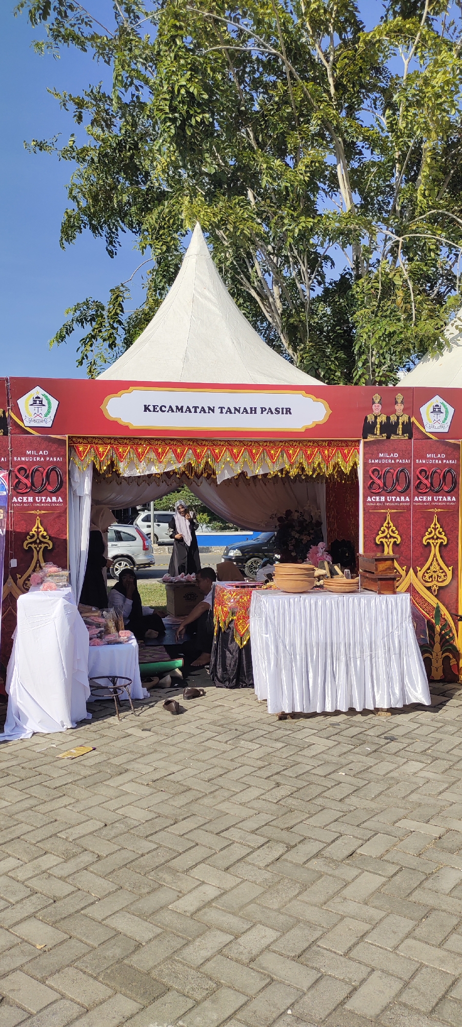 An outdoor event is taking place under a large white tent with red and gold trim. The tent is set up on a paved area with a herringbone brick pattern. In the background, trees with green leaves are visible against a clear blue sky. Several people are present, some standing and some sitting. One woman in a black hijab and abaya is standing and gesturing towards the viewer. Another woman in a light-colored hijab and abaya is sitting behind a table covered with a white cloth, where some pink wrapped items are displayed. A man in a white t-shirt is sitting on the ground. In front of the tent, two tables are set up. One table, on the left, is covered with a white cloth and displays pink wrapped items. The other table, on the right, is also covered with a white cloth and displays wooden bowls and boxes. The tent has signage that reads "KECAMATAN TANAH PASIR". On the right side of the tent, there are vertical panels with images of two men in formal attire and text that reads "MILAD SAMUDERA PASAI 800 ACEH UTARA". On the left side of the tent, similar panels are visible. Cars are parked behind the tent. The overall scene suggests a cultural event or festival. The location is identified as Meunasahaluedrienlhoksukon Barat, Indonesia.