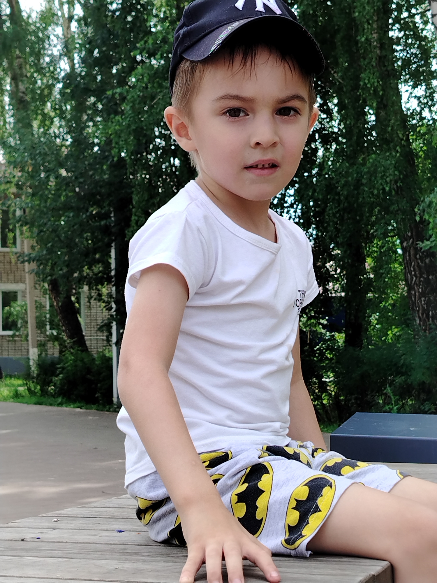 A young boy wearing a white t-shirt, gray shorts with a Batman logo pattern, and a black baseball cap sits on a wooden bench outdoors. The boy is looking directly at the camera with a neutral expression, and his left arm is resting on his leg. Behind him, lush green trees and foliage create a natural backdrop, with a building visible on the left side of the frame. The lighting suggests it is daytime, and the setting appears to be a park or a similar outdoor recreational area. The boy's shorts have a repeating pattern of yellow Batman logos.