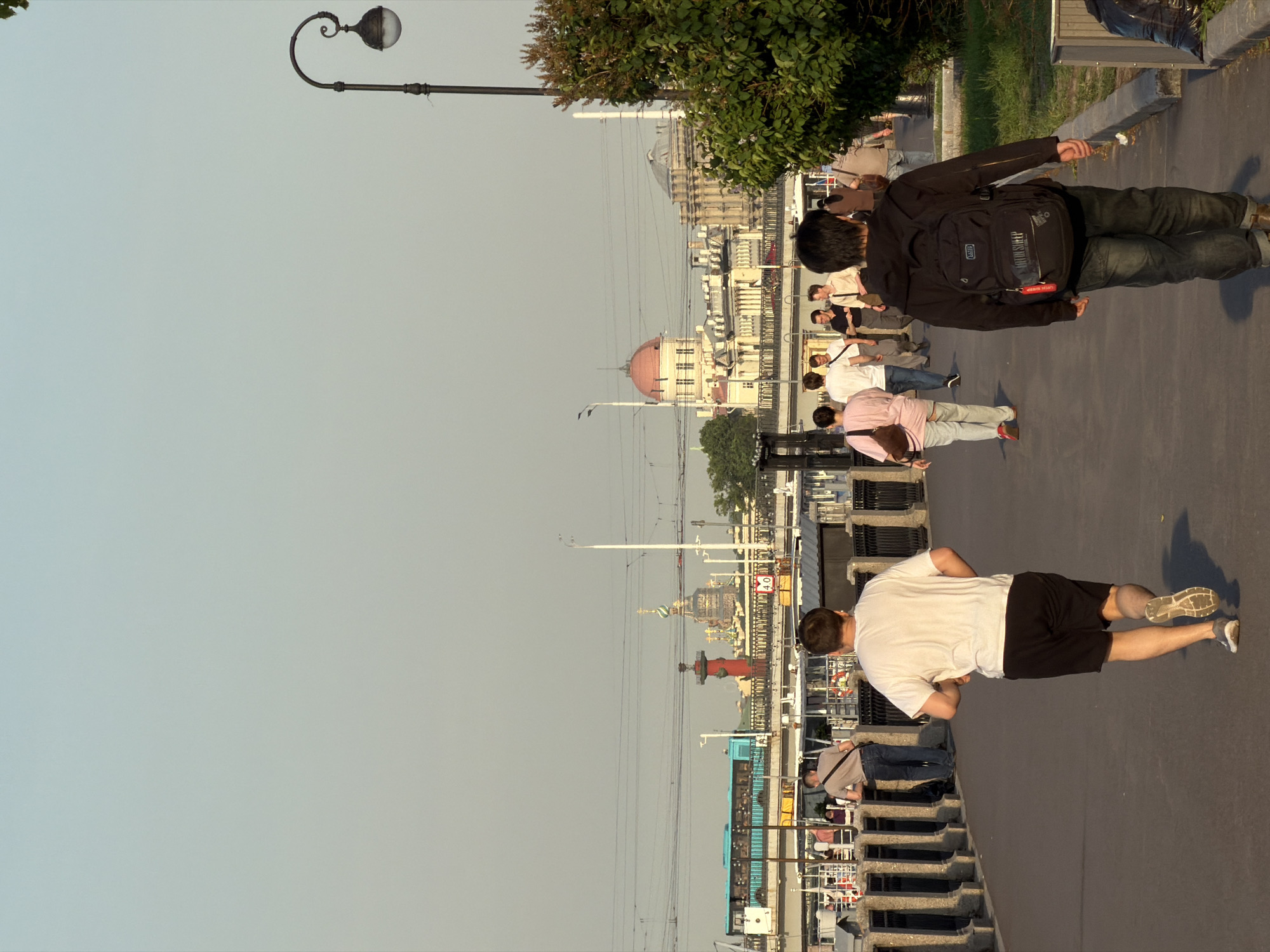 The image depicts a sunny day along the embankment of the Neva River in Saint Petersburg, Russia. In the foreground, a man in athletic shorts and a t-shirt runs away from the camera. Further down the paved walkway, a group of people strolls, some engaged in conversation. In the background, a light blue bus crosses a bridge, and various buildings, including a domed structure, are visible. A street lamp with an ornate design stands to the right, and lush green bushes line the embankment. The scene suggests a casual, outdoor activity in a bustling urban environment.