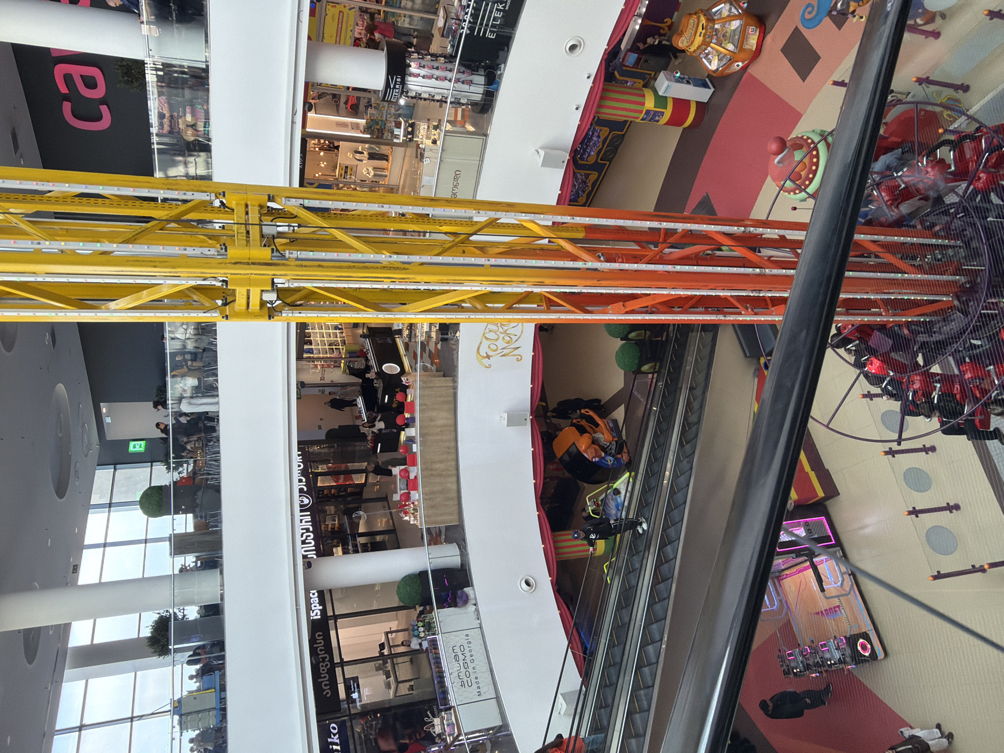 The image depicts the interior of a multi-story shopping mall, potentially in Tbilisi, Georgia, given the presence of Georgian script on shop signs. The perspective is a high-angle shot, possibly from an upper level, looking down. The foreground is dominated by the structural supports of an amusement ride, with yellow and orange metal lattice beams crisscrossing the view. In the background, one can see various retail stores across different levels, escalators transporting people, and a circular amusement ride with rotating seats. People are seen shopping and moving through the mall. There are also games and attractions, including a claw machine game and what appears to be a game with hanging seats. The overall environment is bright and modern.