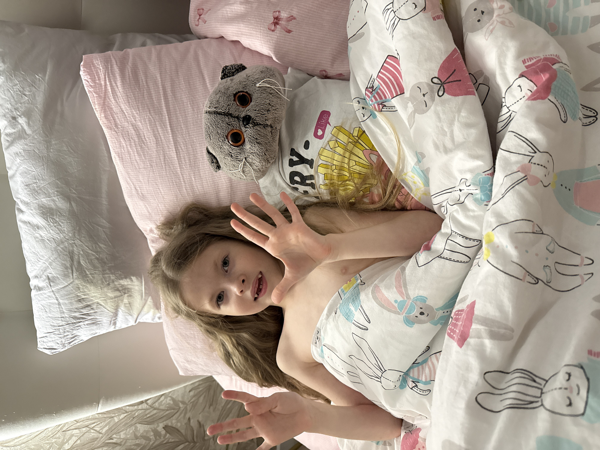 A young girl lies in a bed with white, pink, and patterned bedding. She has long, wavy blonde hair and is reaching towards the camera with both hands, her expression indicating she is awake and engaged. Beside her is a gray stuffed cat wearing a white t-shirt with a french fries design. The bedding features various cartoon-like drawings. A white pillow is visible in the background. No details suggest that the image was taken in the city of Izhevsk, Russia.