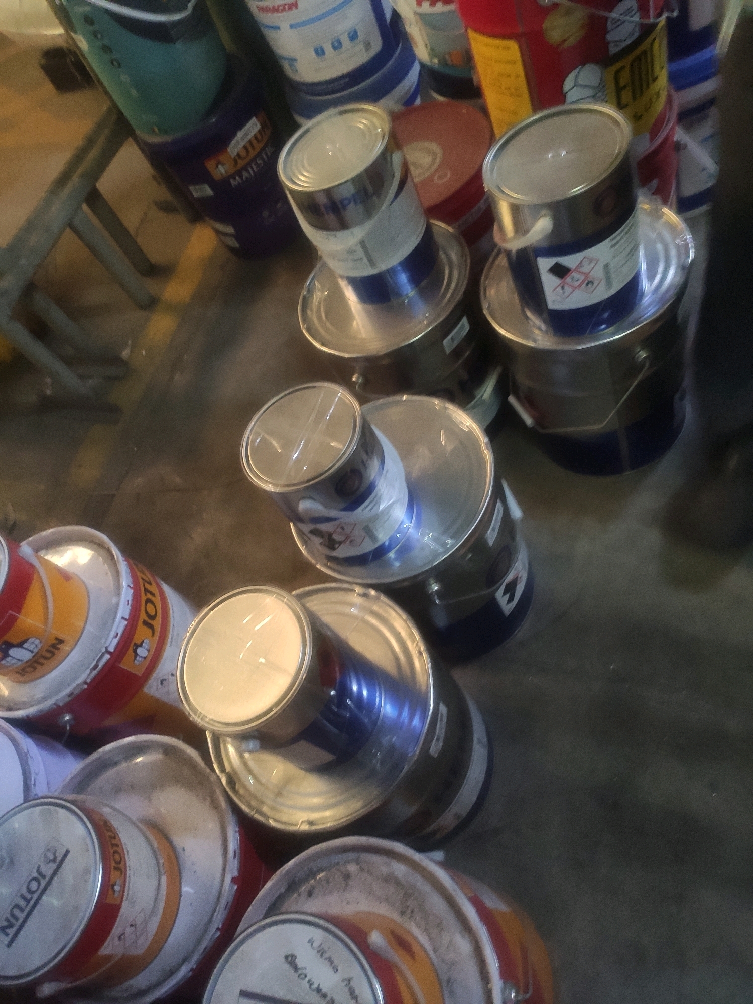 The image shows a collection of paint cans stacked on a floor in what appears to be a storage area or workshop. The cans are of various sizes and brands, including Jotun and Majestic. Some cans are open, revealing paint inside, while others are sealed. The floor is concrete and appears to be slightly dusty. There are no people visible in the image. The lighting is dim, suggesting it may be indoors or late in the day. The scene is cluttered with the paint cans, suggesting a work-in-progress or a stock of supplies.