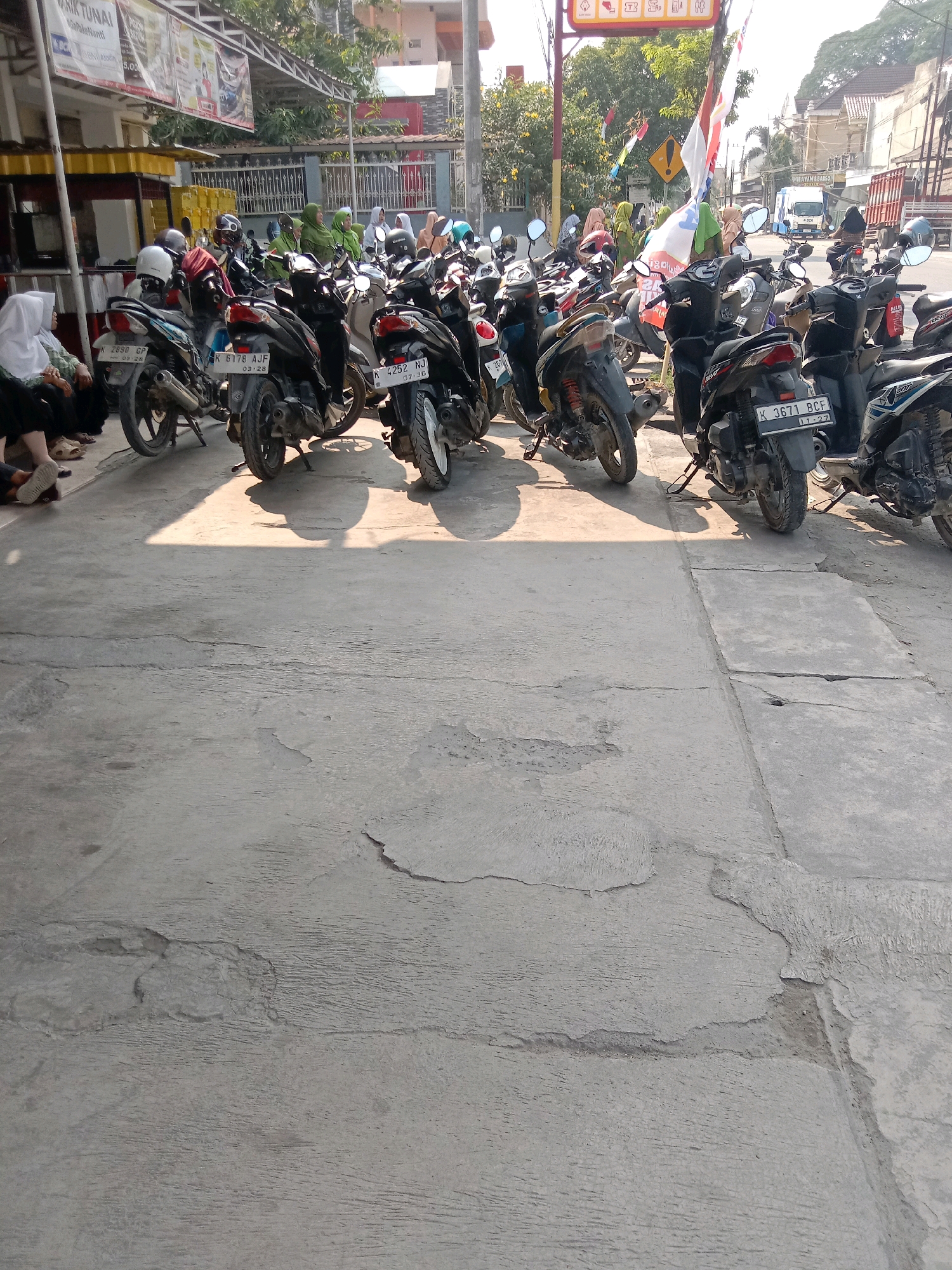 The image depicts a street scene in Kalisari, Indonesia, featuring a parked line of motorcycles. Several individuals are visible, some seated and others standing near the motorcycles. The environment suggests a commercial or public area with buildings and trees in the background, and a truck is visible on the road. The overall impression is a snapshot of everyday life in this Indonesian city.