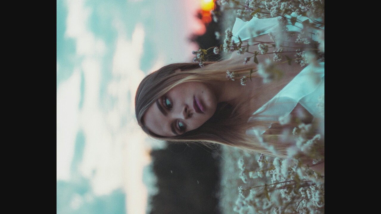A medium shot captures a young woman with blonde hair and blue eyes in a field of white flowers. She is wearing a white dress with a flowing neckline and is looking off to the side with a thoughtful expression. The background features a blurry landscape with trees and a sunset in the distance. The sky is a mix of blue and pink clouds. The lighting is soft and diffused, creating a dreamy and serene atmosphere. There are two small, dark insects in the air near the woman's head. The overall mood of the image is peaceful and contemplative.