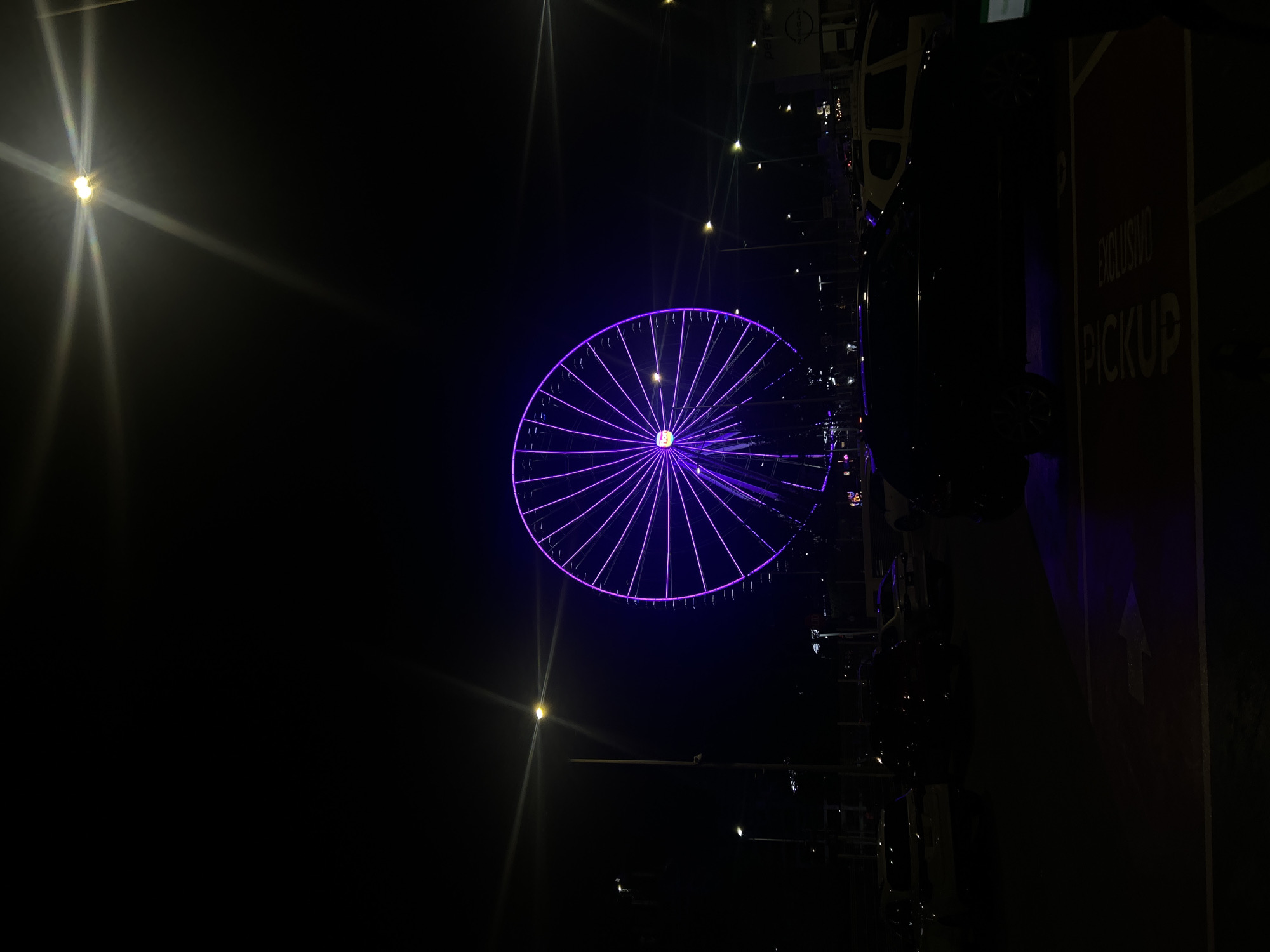 The image depicts a night scene in what appears to be a public space. A large Ferris wheel illuminated with purple lights is the focal point. The wheel's design consists of radiating spokes connecting to the central hub. To the right of the wheel, a partially visible sign with the words "EXCLUSIVO PICKUP" and an upward-pointing arrow can be seen. Streetlights emit a bright glow with noticeable lens flares. The surrounding environment is dark, with other structures and objects only discernible as silhouettes. There is no clear indication of the exact location within San Bernardino Tlaxcalancingo, Mexico, or the presence of people or specific actions.