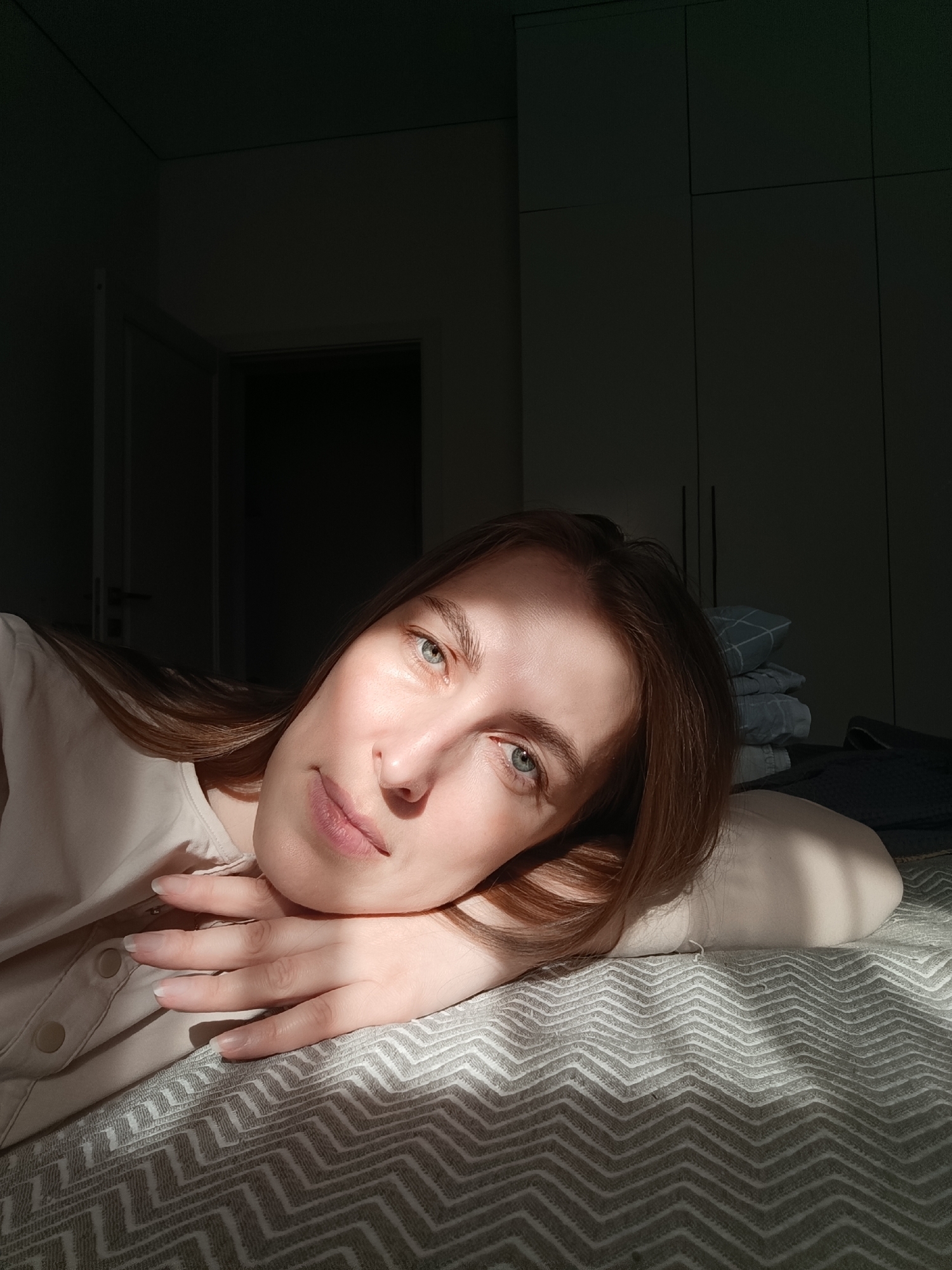 A fair-skinned woman with light brown hair and blue eyes is lying on a bed, resting her head on her hand. She is wearing a light-colored, buttoned shirt. The bed has a gray patterned cover. A stack of folded linens is visible in the background. The light source appears to be coming from the right, casting shadows on the wall and the woman's face. An open doorway is visible to the left. The scene appears to be indoors, possibly in a bedroom. The location is Chaikovsky, Russia.
