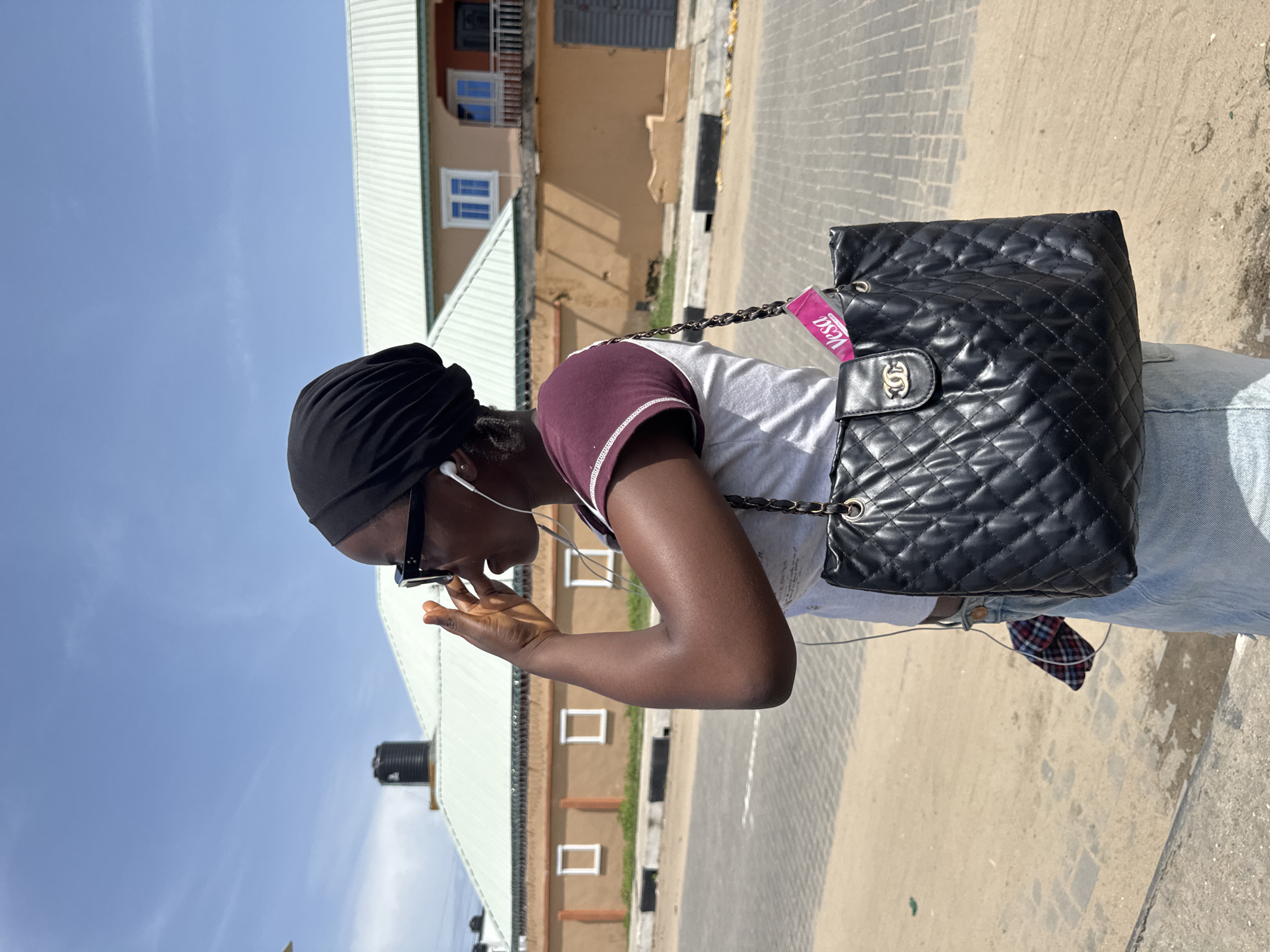A young, Black woman with dark skin is standing on a paved street. She is wearing a black head covering, sunglasses, a white and maroon t-shirt, and light blue jeans. She has earbuds in her ears and is holding her left hand near her face, as if she is adjusting her sunglasses or talking on the phone. A large, black, quilted purse with a chain strap is slung over her shoulder. A pink tag is attached to the purse. In the background, there are buildings with light brown facades and light green roofs. The sky is blue. The location is possibly in the city of Badagry, Nigeria.