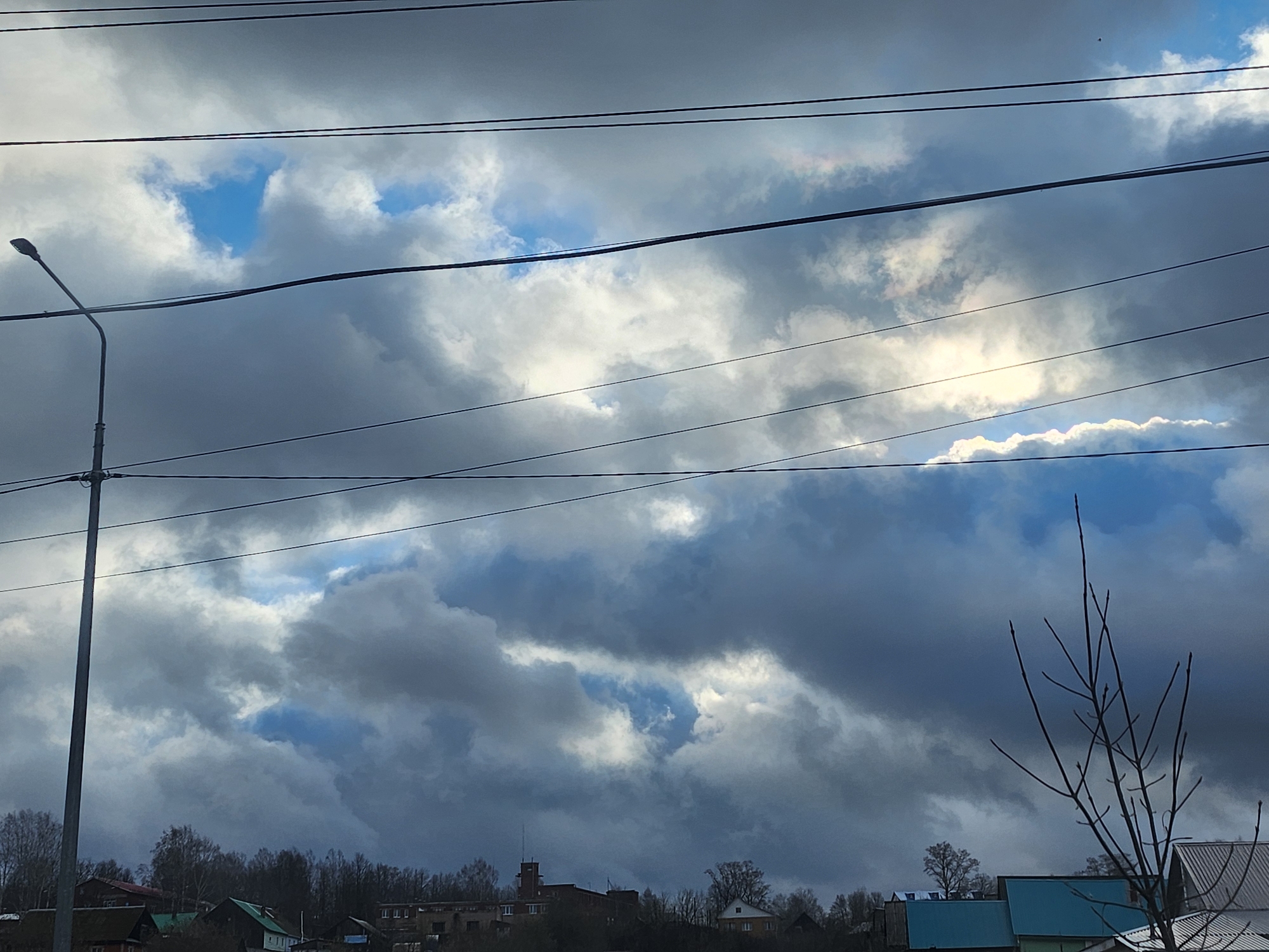 The image captures an expansive, dynamic skyscape over a semi-urban or rural settlement in Krasnogorskoye, Russia. The sky is largely covered by dense, dark grey and white cumulus clouds, with bright patches of sunlight breaking through and illuminating certain cloud sections. Several distinct areas of clear blue sky are visible where the clouds part.
Multiple black utility wires or power lines stretch horizontally across the upper and middle portions of the frame, creating linear elements against the sky. On the far left, a tall, grey streetlamp with a curved arm and a light fixture extends upwards, partially supporting some of these wires.
Below the dramatic sky, the upper parts and roofs of various buildings and houses are discernible. These structures appear to be clustered, with some having dark roofs and one prominent building in the center-left featuring a distinctive, possibly older, brick-like tower. A line of bare, leafless trees, typical of late autumn, winter, or early spring, forms a horizon line between the buildings and the sky. On the far right, the stark, upward-reaching branches of a bare tree or large bush are silhouetted against the sky.
No specific activity or event is taking place; the image is a static depiction of the environment. The time of day appears to be daytime, likely mid-morning or afternoon, given the intensity of the light filtering through the clouds. The weather is overcast but with glimpses of brighter conditions, suggesting changeable skies. There is no visible text in the image.