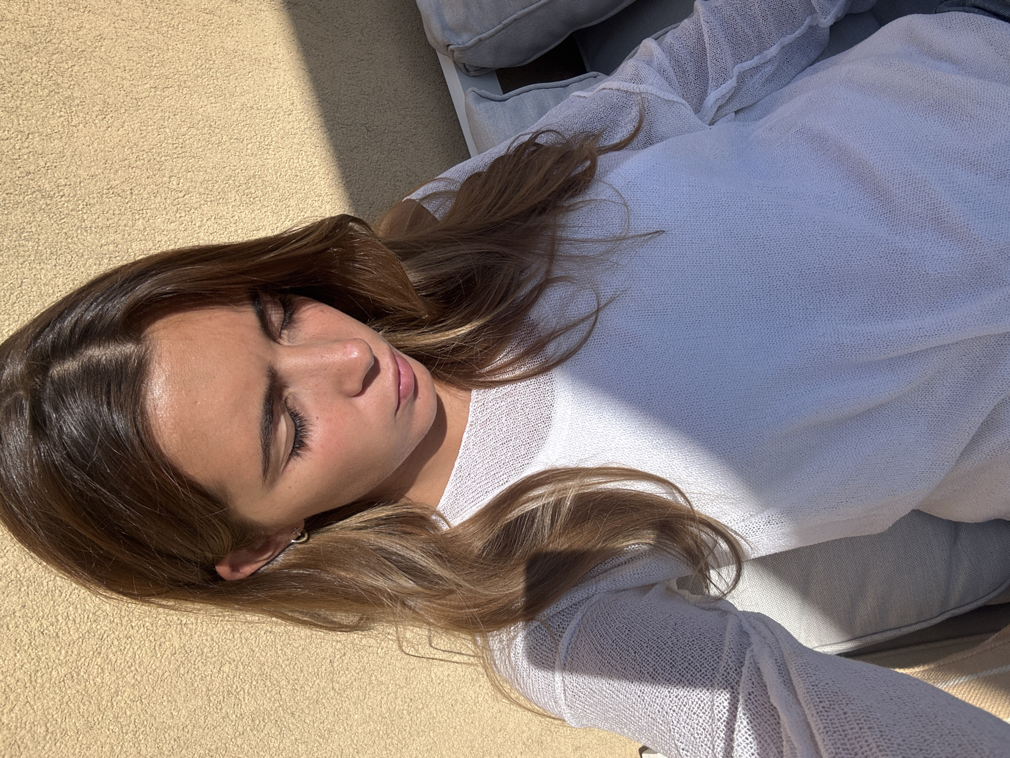 A young woman with long, medium brown hair is depicted resting outdoors under bright sunlight. She is lying on her back or side, with her eyes closed, suggesting a state of deep relaxation or sleep. She wears a white, loosely knitted long-sleeved top. Her skin shows a natural tan, highlighted by the direct sun.
The setting appears to be an outdoor space, possibly a patio, balcony, or garden, in Vila Nova de Gaia, Portugal. On the left side of the frame, there is a light-colored, sandy-textured ground or outdoor carpet. To the right, she is resting against light grey outdoor cushions, which appear to be part of a sofa or chaise lounge.
The scene is bathed in bright daylight, casting strong, distinct shadows across her body, hair, and the surrounding surfaces. This indicates a clear, sunny day, likely during the mid-morning or afternoon hours. The overall mood is peaceful and serene. There is no visible text in the image.