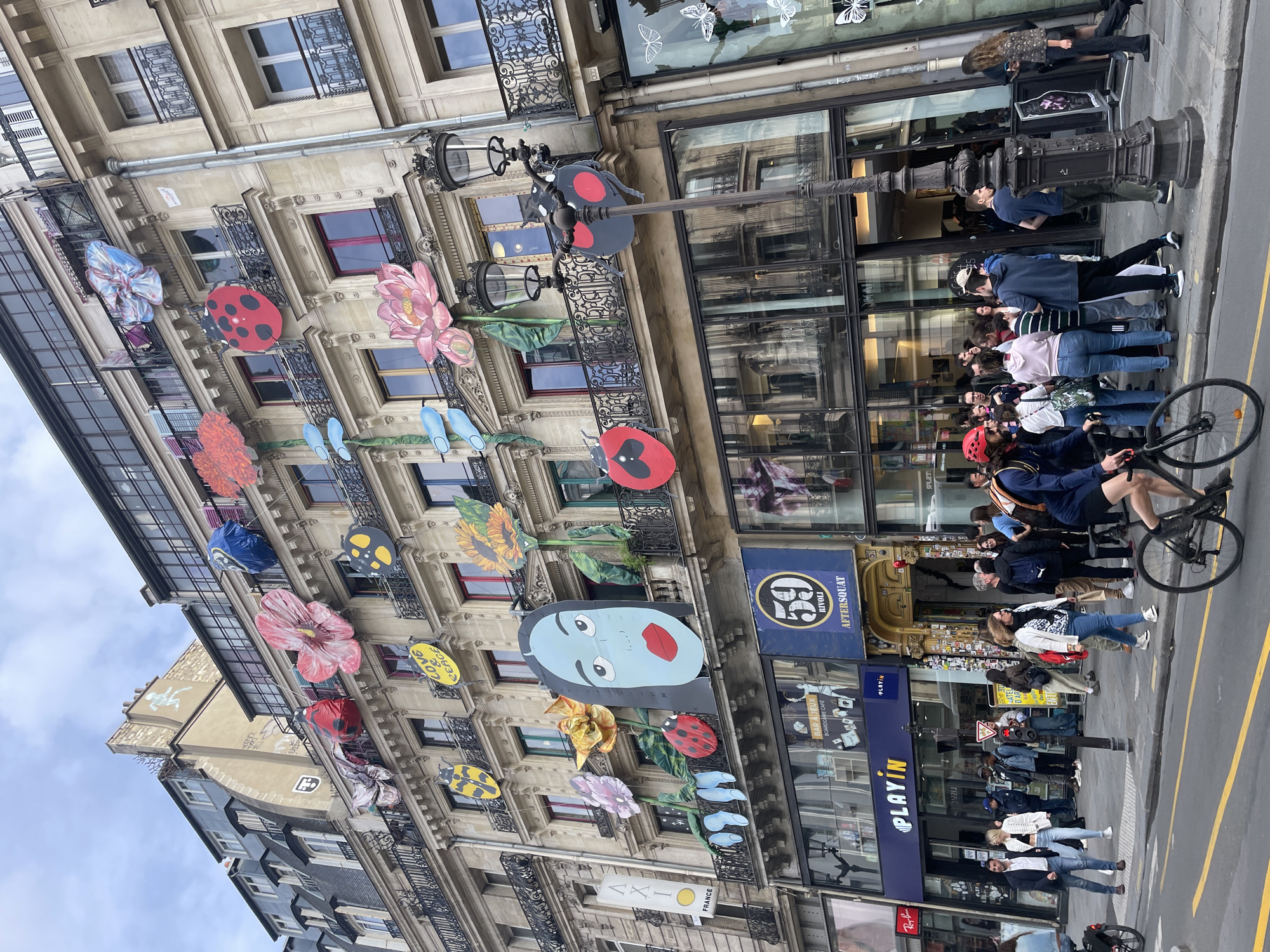The image captures a street scene in Paris, France, with a focus on the facade of a multi-story building adorned with large, colorful art installations. The artwork includes oversized flowers, ladybugs, and a stylized face with blue skin and red lips. These elements are suspended from balconies and walls, creating a vibrant and whimsical display. Below the decorated building, shops are visible, including "PLAYIN" and "59 RIVOLI AFTERSQUAT". A crowd of people is gathered on the sidewalk in front of these establishments, suggesting a public space with ongoing activity. Some individuals are walking, others are standing in groups, and one person is riding a bicycle in the foreground. The architecture of the building is typical of Parisian streetscapes, with ornate detailing and balconies. The sky is overcast, casting a diffused light on the scene.