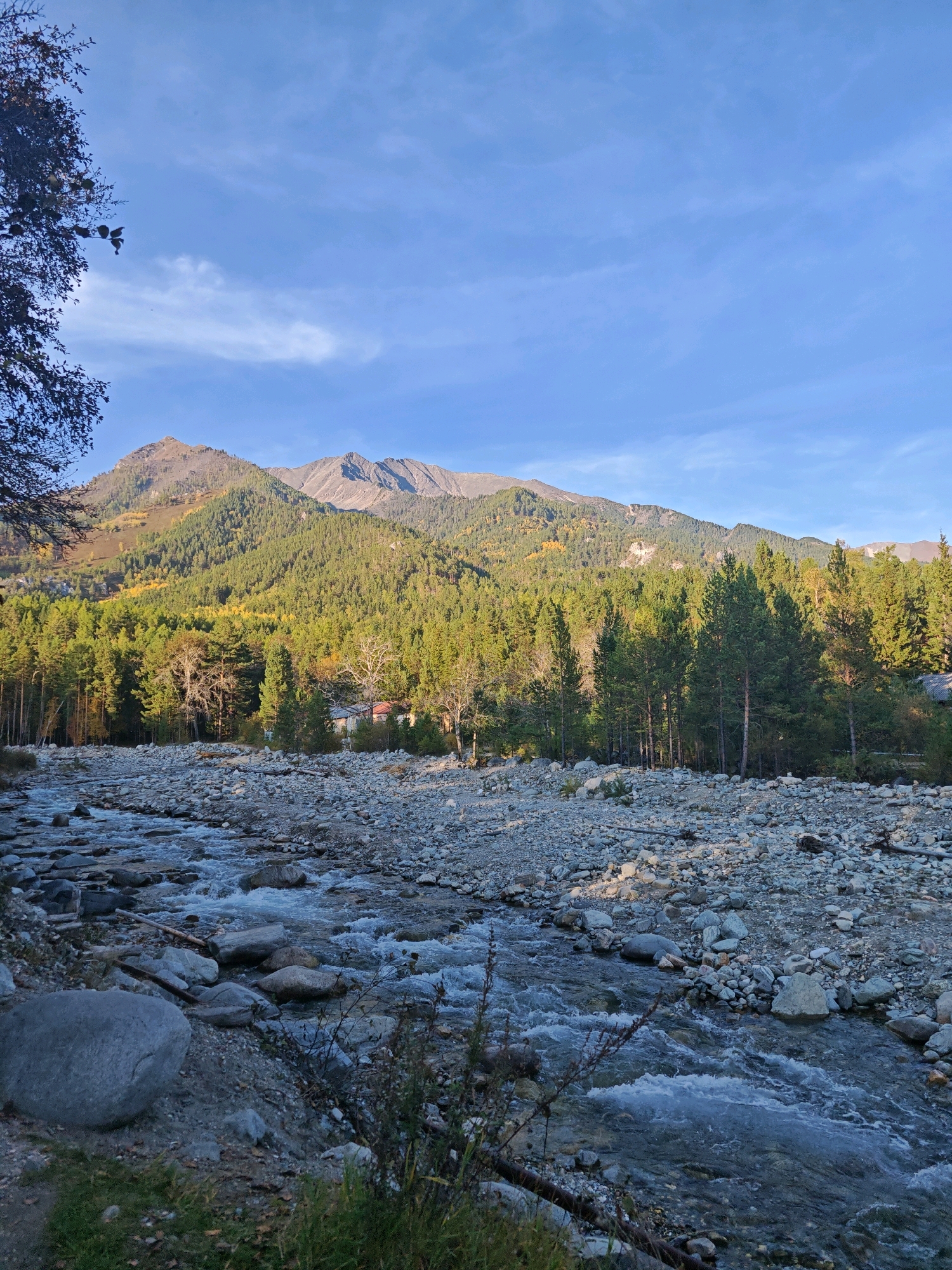 The image depicts a scenic view of a mountain landscape near the city of Arshan, Russia. In the foreground, a rocky riverbed is visible with a stream of clear water flowing over smooth stones. The river banks are lined with various sized rocks and some sparse vegetation. Beyond the river, a dense forest of evergreen trees covers the lower slopes of the mountains. Patches of yellow and green foliage suggest it might be autumn. In the middle ground, a rustic wooden building with a red roof is nestled amongst the trees. The background is dominated by a majestic mountain range, with its rugged peaks stretching across the horizon under a clear blue sky with a few wispy white clouds. The overall scene conveys a sense of natural tranquility and wilderness.