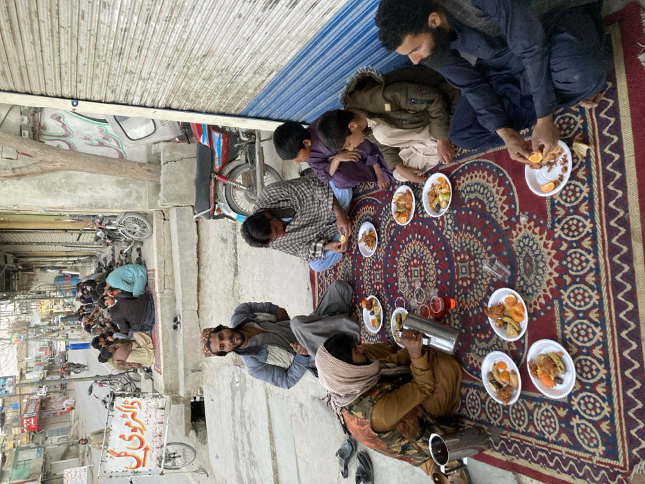The image depicts an outdoor communal meal on a paved street or alleyway in Quetta City Tehsil, Pakistan.
In the foreground, six males, consisting of men and boys, are seated cross-legged on a rectangular, patterned rug with red, blue, and gold motifs. They are engaged in eating. Eight small, white, round plates are distributed on the rug, each containing portions of fried food items and cut fruit, including orange segments. A man on the far left, wearing a tan shalwar kameez and a beige headscarf, pours a red beverage from a silver thermos into a clear plastic cup. Another silver jug is nearby. To his right, a man in a blue jacket and grey shalwar kameez sits facing the camera. Further right, a man in a plaid shawl peels an orange over a plate. Two boys, one in purple and one in a beige jacket, sit next to him, observing the food. On the far right, a man with a dark beard and dark blue shalwar kameez is also peeling an orange over a plate. Several pairs of dark shoes are placed on the concrete around the rug.
In the midground, directly behind the main group, a light blue and white motorcycle is parked. A small tree with bare branches grows from a concrete planter. To the left, another group of approximately 10-15 men are seated on elevated concrete steps, also on rugs, appearing to socialize or eat. A white, wheeled sign displaying Urdu script "ڈاکٹر مولوی گل" (Dr. Maulvi Gul) and "پٹاخے" (Patakhe - firecrackers) is prominent on the left side of the street.
The background shows a street receding into the distance, lined with multi-story buildings that appear to be shops, many featuring metal roll-down shutters. Several other motorcycles are parked along the street. A few individuals are visible in the distance, walking or cycling. Utility poles and power lines crisscross overhead. The sky is bright but appears overcast or at dusk.