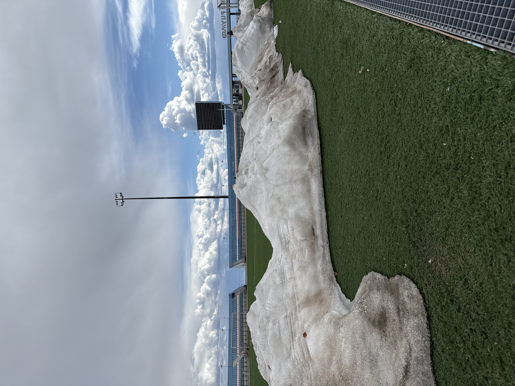 The image depicts a partially melted pile of snow on a sports field, likely artificial turf, adjacent to stadium seating. The sky is partly cloudy, with a mix of dark and bright clouds. A tall lighting tower is visible in the background. The word "BRAV" is visible on a sign, horizontally aligned, above the seating. The location could be the "Arena Druzhba" stadium in Mirny, Russia, based on the presence of the "BRAV" sign and stadium structures visible, which are indicative of sports complexes in the area.