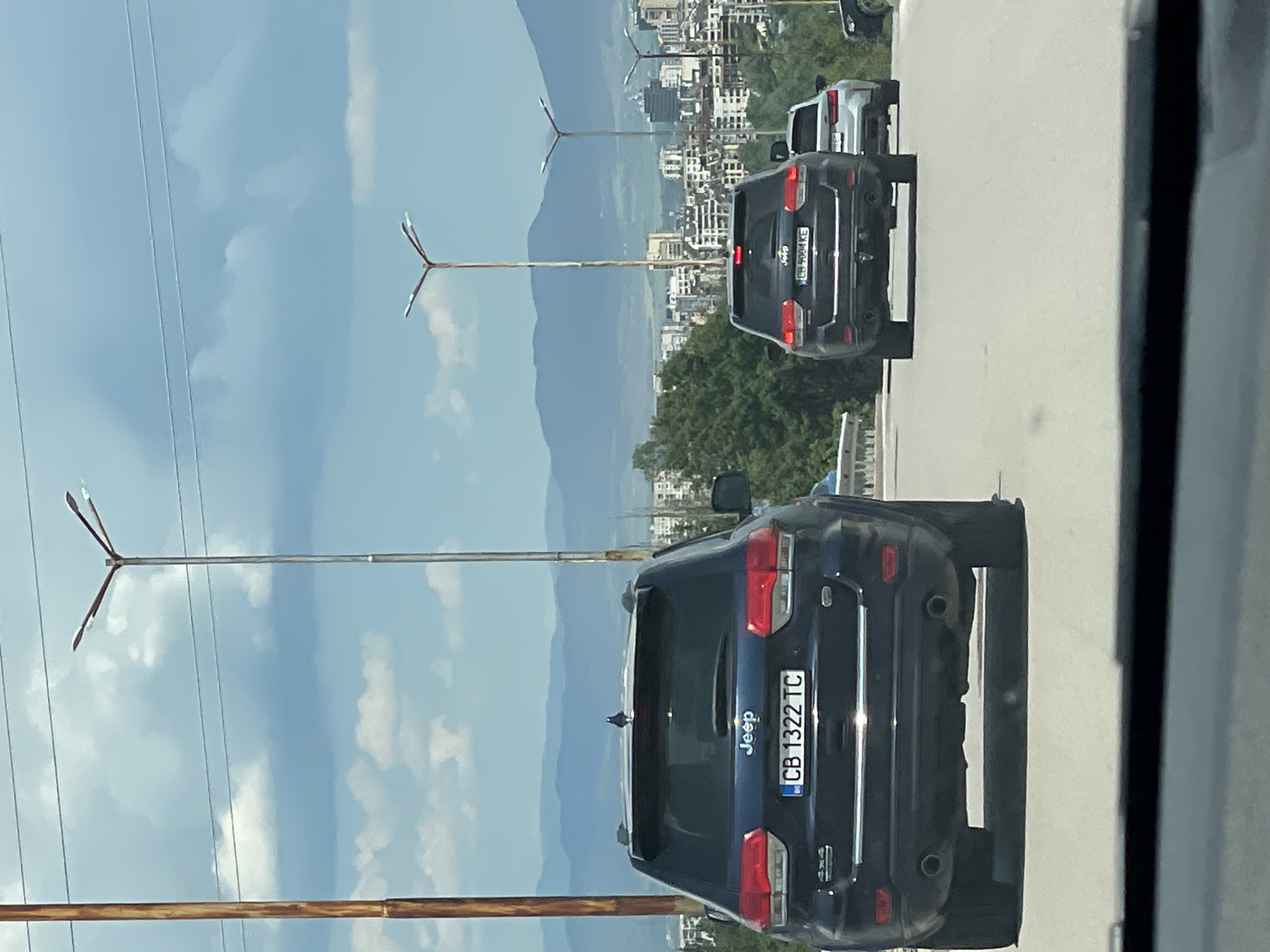 An eye-level, medium shot captures traffic on a highway in Sofia, Bulgaria. In the foreground, the rear of a dark blue Jeep Grand Cherokee is visible, its license plate reading "CB 1322 TC". The vehicle is traveling on a multi-lane road. Further down the highway, another dark Jeep Grand Cherokee with the license plate "CB 9004 KE" is also visible, along with a lighter-colored vehicle behind it. In the background, a cityscape unfolds with numerous buildings and distant mountains, all under a partly cloudy sky. Tall streetlights line the highway. The overall scene depicts a typical urban traffic situation.