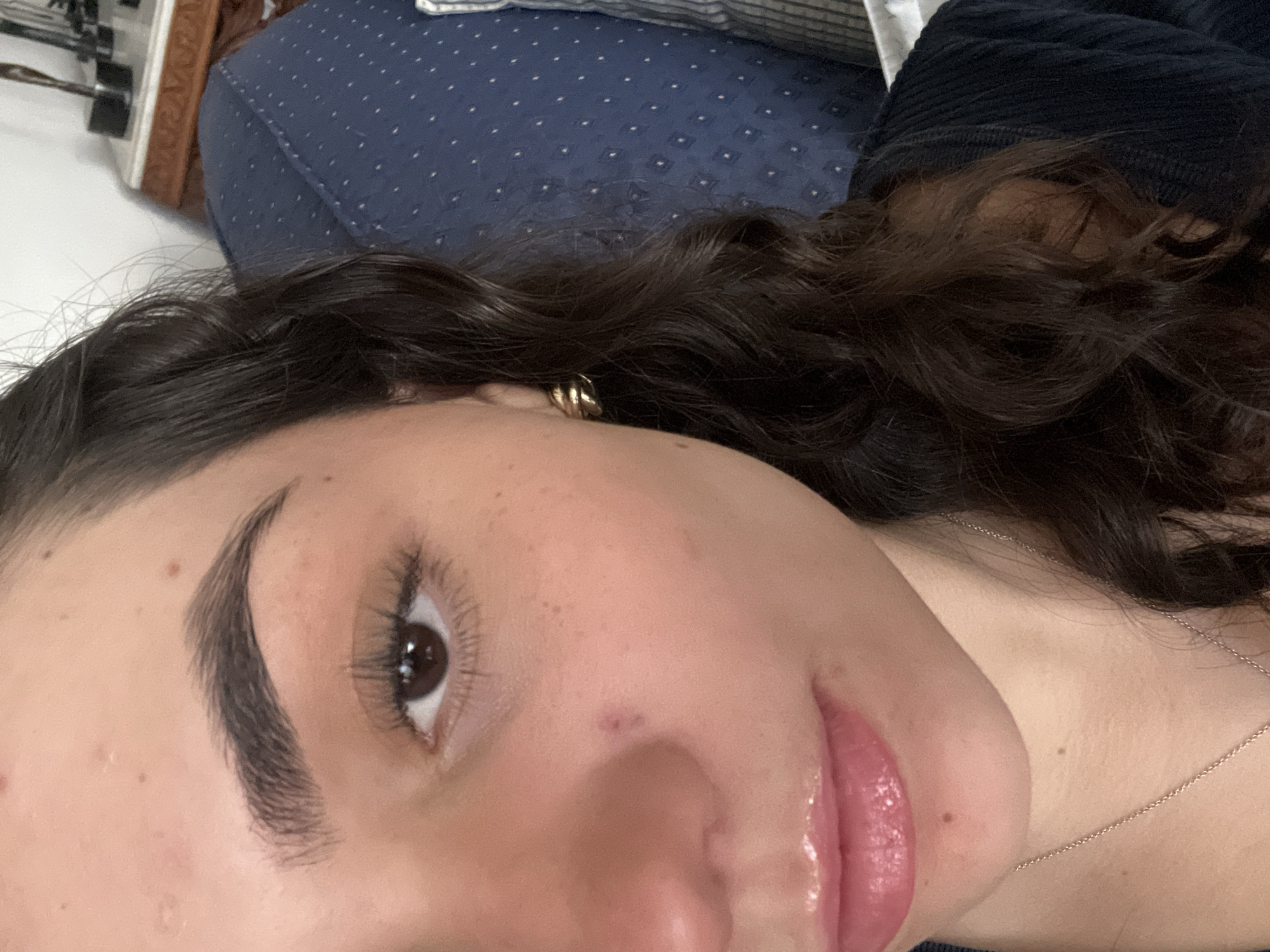 The image depicts a close-up view of a person's face, likely female, featuring dark, curly hair and fair skin. The person has a prominent eyebrow, a brown eye with visible lashes, and full lips with a glossy finish. A gold earring is visible in the ear. A thin silver necklace is also visible. The background is a mix of textures, including a blue, patterned fabric (possibly upholstery) and a black, corduroy-like material, indicating an indoor setting. The person is lying down. The location is unknown.