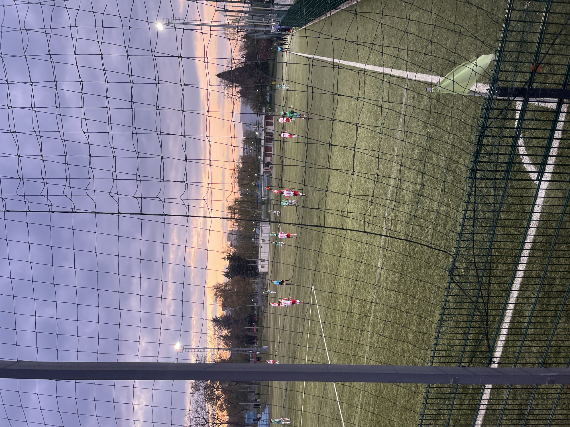 The image captures an outdoor soccer field at dusk or early evening, viewed through a prominent, dark netting in the foreground. The scene is oriented vertically, but depicts a landscape view of the pitch.
In the midground, an artificial green turf field is visible, marked with white lines indicating boundaries and possibly penalty areas. Numerous soccer players are scattered across the field, some wearing red and white uniforms, others in green. A referee in a blue shirt is also present. A bright green corner flag stands near the right sideline. Behind the foreground netting and to the right, there's another, larger dark green metal fence structure.
The background features a colorful sky, displaying soft gradients of purple, pink, and orange hues, characteristic of sunset or twilight. Buildings and trees are discernible in the distance beneath the sky. Two bright floodlights on tall poles are active, illuminating parts of the field and standing out against the fading daylight. The lighting suggests an evening match or training session.
No visible text is identifiable in the image.