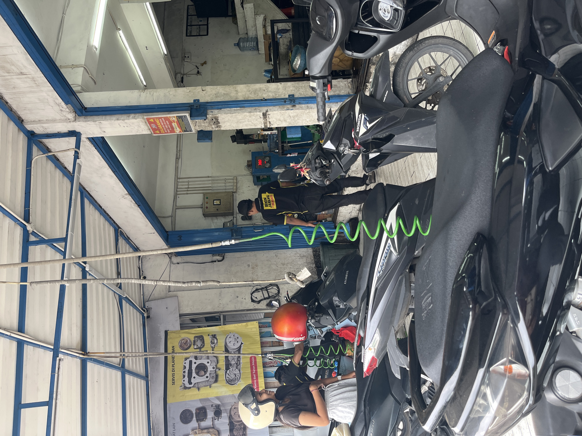 The image shows an outdoor motorcycle repair shop in Denpasar, Indonesia. The shop appears to be located under a covered structure with a white roof supported by blue beams. Several motorcycles are parked in the foreground, with their seats visible. In the middle ground, two individuals are present. A woman wearing a white helmet and a dark t-shirt with a grey skirt is smiling and looking towards the right. Next to her, a person in a black shirt with yellow lettering and black pants is holding a red motorcycle helmet. Behind them, a man in a similar black shirt and pants is walking away from the camera towards the back of the shop. The background reveals the interior of the shop, with tools, equipment, and a water cooler visible. A yellow banner with images of engine parts hangs on the left wall, and a red and yellow warning sign is posted on a pillar to the right. The overall scene depicts a typical day at a motorcycle service center in Denpasar.