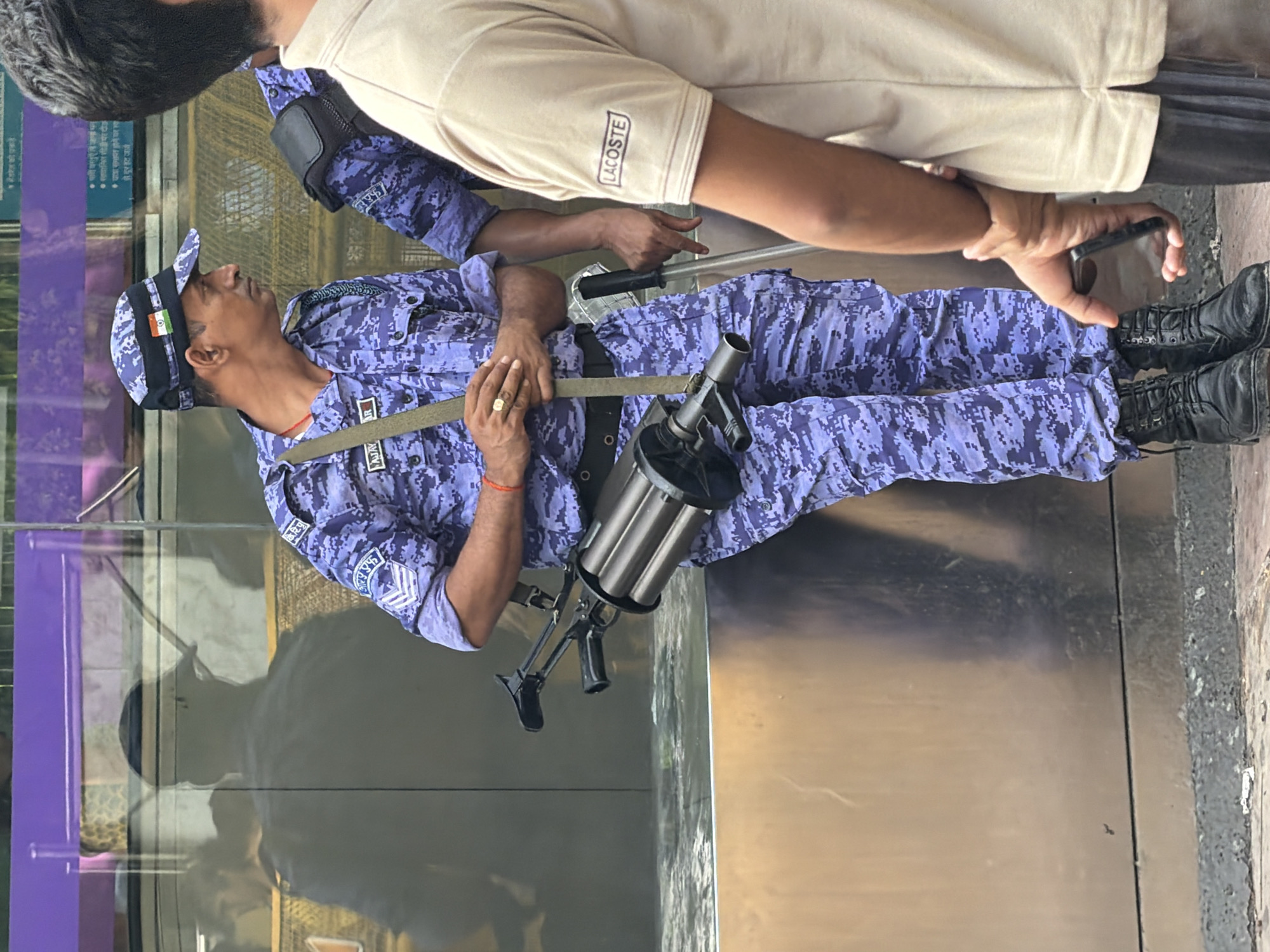 In New Delhi, India, a male police officer in a blue camouflage uniform with an Indian flag patch on his cap stands next to another uniformed officer. The primary officer holds a multi-barreled firearm with a black grip. He wears a gold ring on his right hand and a red thread around his wrist. His uniform has patches with text, including "AJAY" and a rank insignia on the sleeves. The other officer is partially visible, holding a baton. In the foreground, a person in a light-colored t-shirt with a "LACOSTE" logo is seen from the chest down, holding a smartphone. The background features a reflective glass wall with purple accents, showing a shadow of another person. The scene takes place on a paved area.