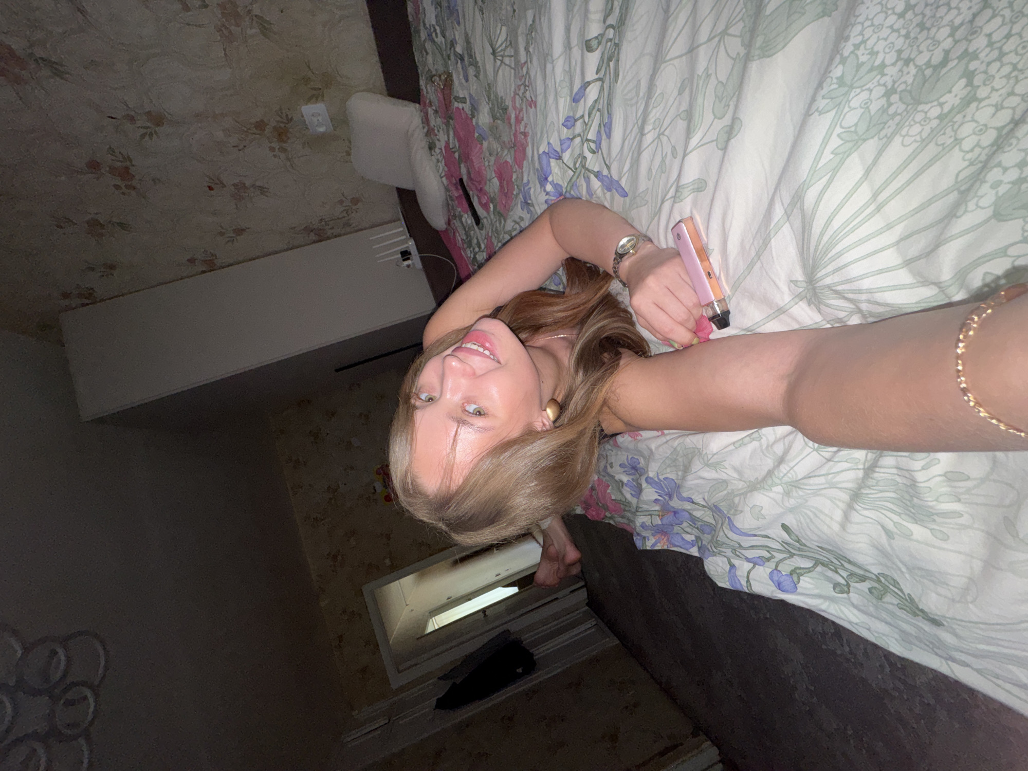 A young woman with blonde hair and light green eyes is lying on a bed in a room, looking towards the camera and smiling. She is wearing large gold hoop earrings and a watch on her left wrist. Her left arm is extended towards the camera, holding a pink electronic cigarette device. The bed is covered with a floral-patterned duvet. In the background, there is a doorway leading to another room, where a pair of feet can be seen. The room appears to be somewhat dilapidated, with peeling wallpaper. The lighting is dim, suggesting it is nighttime or the room has no natural light source. The image was taken in Chaikovsky, Russia.