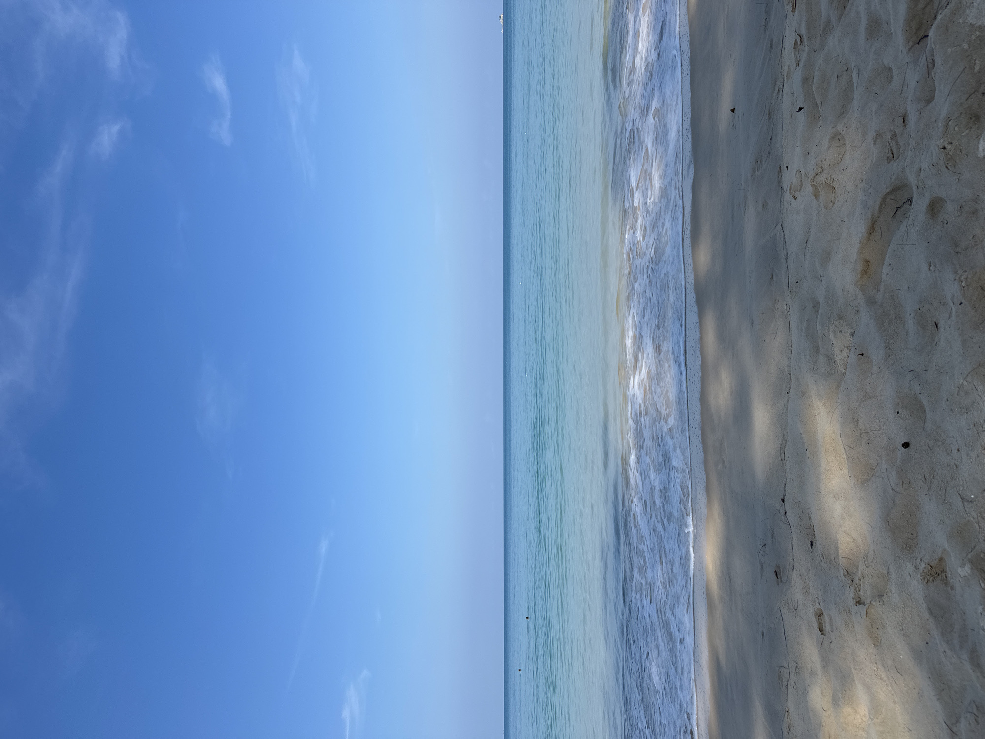The image shows a beach scene in Choeng Thale, Thailand. On the left is a clear blue sky with wispy clouds. In the center, turquoise water meets the shore with foamy waves. On the right, light-colored sand displays shadows, possibly from trees.