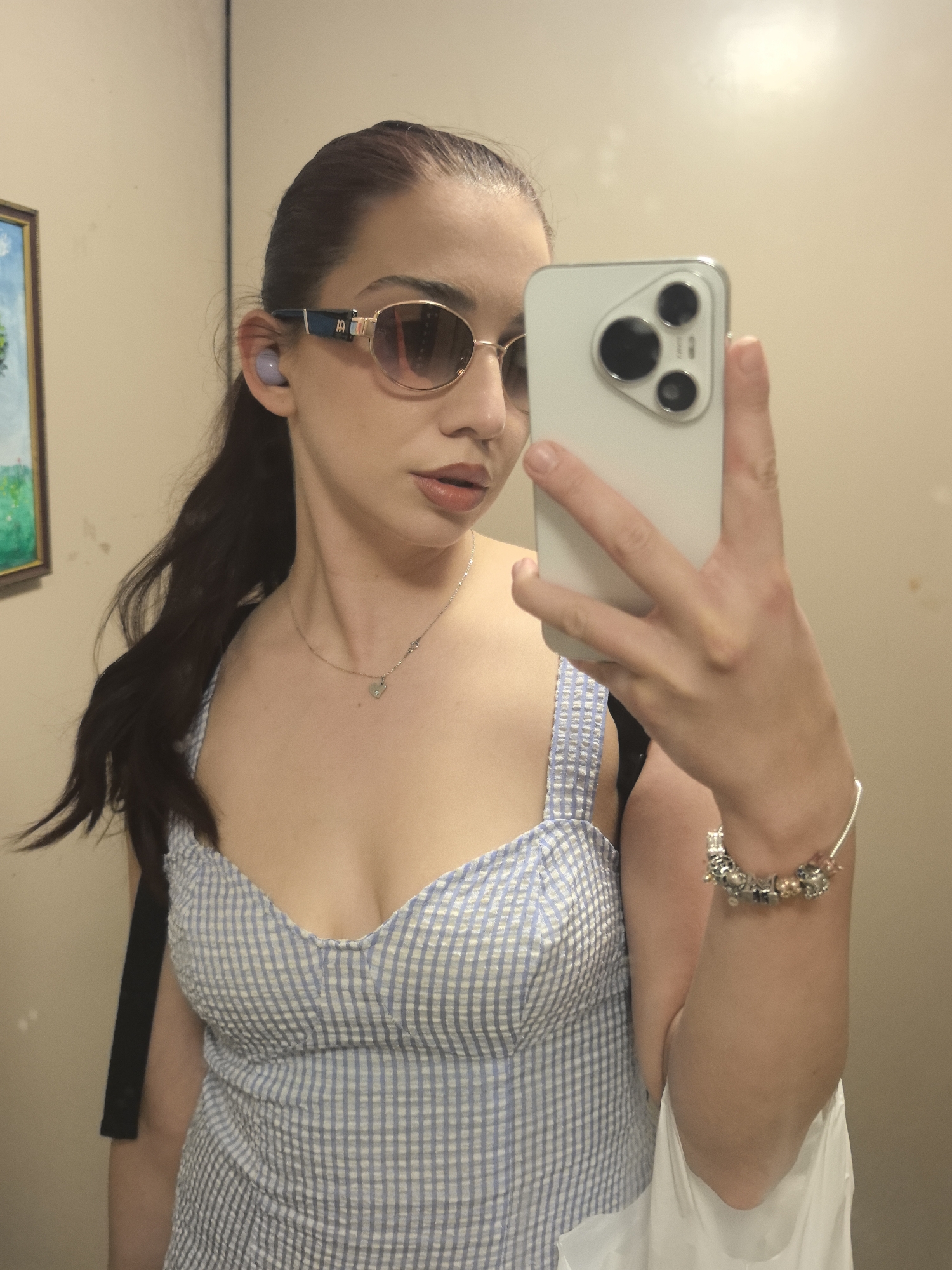 A woman with brown hair pulled back into a ponytail is taking a selfie using a smartphone. She is wearing round sunglasses with a gold frame and blue accents on the sides, and a small lavender earbud is visible in her left ear. A silver necklace with a heart-shaped pendant adorns her neck. She is wearing a blue and white checkered dress with thick straps. A black strap and a white bag are also visible. She is wearing a silver bracelet with several charms. The background appears to be a plain, beige-colored wall. There is a framed picture hanging on the left wall. The location of the image is unknown and not necessarily located in Stara Zagora, Bulgaria.