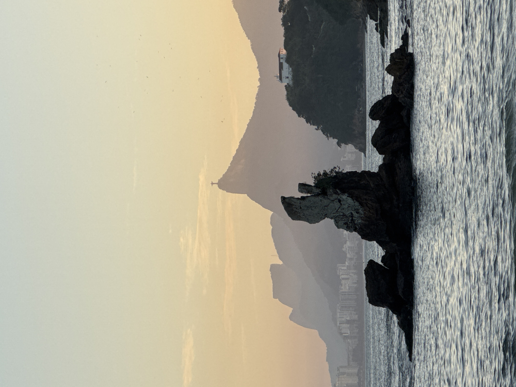 The image depicts a coastal scene with a prominent rock formation in the foreground, surrounded by the ocean. In the background, a city skyline and a series of misty mountains are visible. On the summit of one of the mountains, the iconic Christ the Redeemer statue is discernible. A white building with a red roof is situated on a forested hill to the right of the statue. The sky exhibits a gradient of pale yellow to light blue, with a few small birds scattered across it. The water is calm with gentle ripples. The overall impression is that of a serene sunset or sunrise over a coastal city. This is likely a view of Rio de Janeiro, as Niterói is across the Guanabara Bay from Rio, and the Christ the Redeemer statue is in Rio.