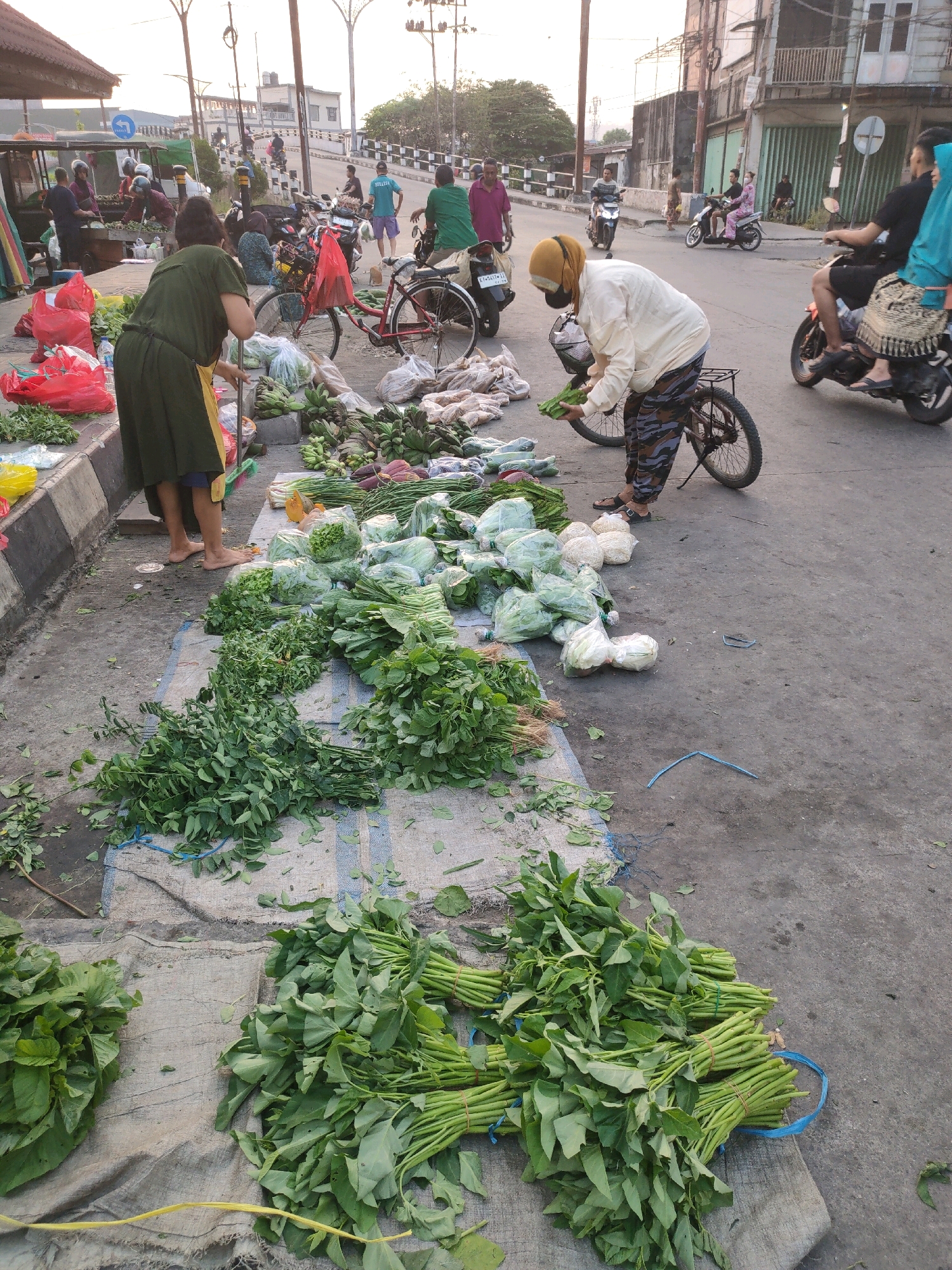 A street market scene is depicted in Samarinda, Indonesia, under a bright, possibly early morning or late afternoon sky. The ground is paved with asphalt, and a concrete curb separates the market area from the road. A variety of fresh produce, primarily leafy green vegetables and some root vegetables, are displayed for sale.
In the foreground, large bundles of green vegetables are arranged on a textured, cloth-like mat. To the left, a person wearing a dark green tunic and a yellow apron is seen from the waist down, standing barefoot and appears to be sweeping or cleaning the area with a broom. Their legs are bare.
Further into the market, more vegetables are laid out, including bunches of what appear to be spinach, long beans, and cabbages wrapped in plastic. Several bags of produce, some red and some white, are also visible.
Several people are present in the scene, some interacting with the market stalls and others in transit. A person in a light-colored headscarf and a cream-colored long-sleeved top is bent over, possibly tending to the produce or interacting with a customer. They are wearing camouflage-patterned pants and sandals, and are near a bicycle with a basket.
In the background, motorbikes are a common mode of transportation, with several individuals riding them. Some are parked, while others are moving along the street. People are also seen walking and standing around. The architecture in the background suggests an urban environment with buildings and streetlights. The overall atmosphere is that of a busy, open-air market.