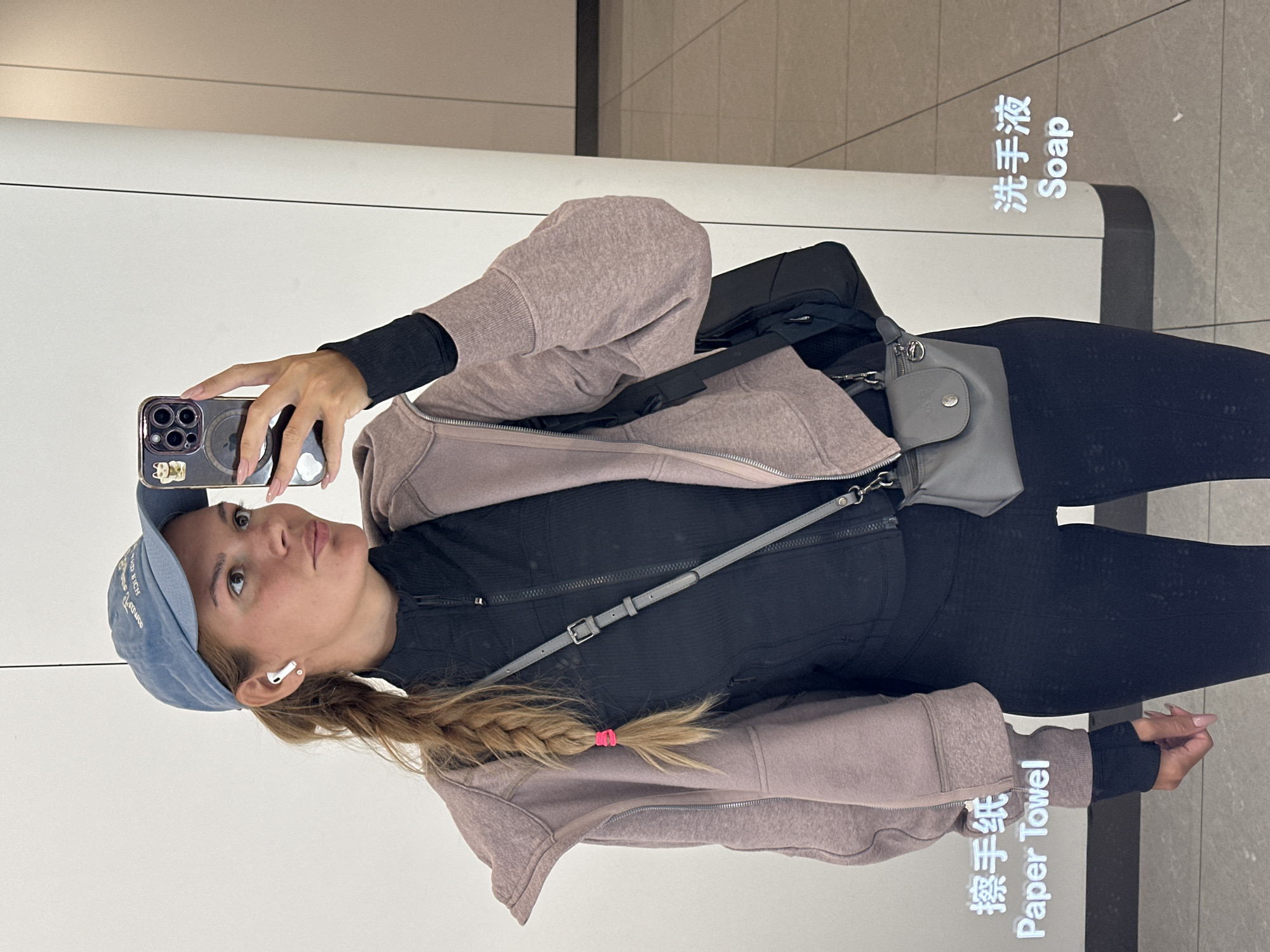 A woman is taking a mirror selfie in what appears to be a public restroom in Jianyang City, China. She is wearing black leggings and a black athletic top, with a mauve-colored hooded jacket. A blue baseball cap is on her head, and she has an earbud in her ear. Her hair is blonde and styled in a braid with a pink hair tie. She is holding a smartphone in her hand, taking the picture. A gray crossbody bag is slung across her body. The background indicates a bathroom setting, with a soap dispenser and paper towel dispenser labeled in both English and Chinese characters. The dispensers appear to be attached to a light-colored wall. The floor is a gray tile.