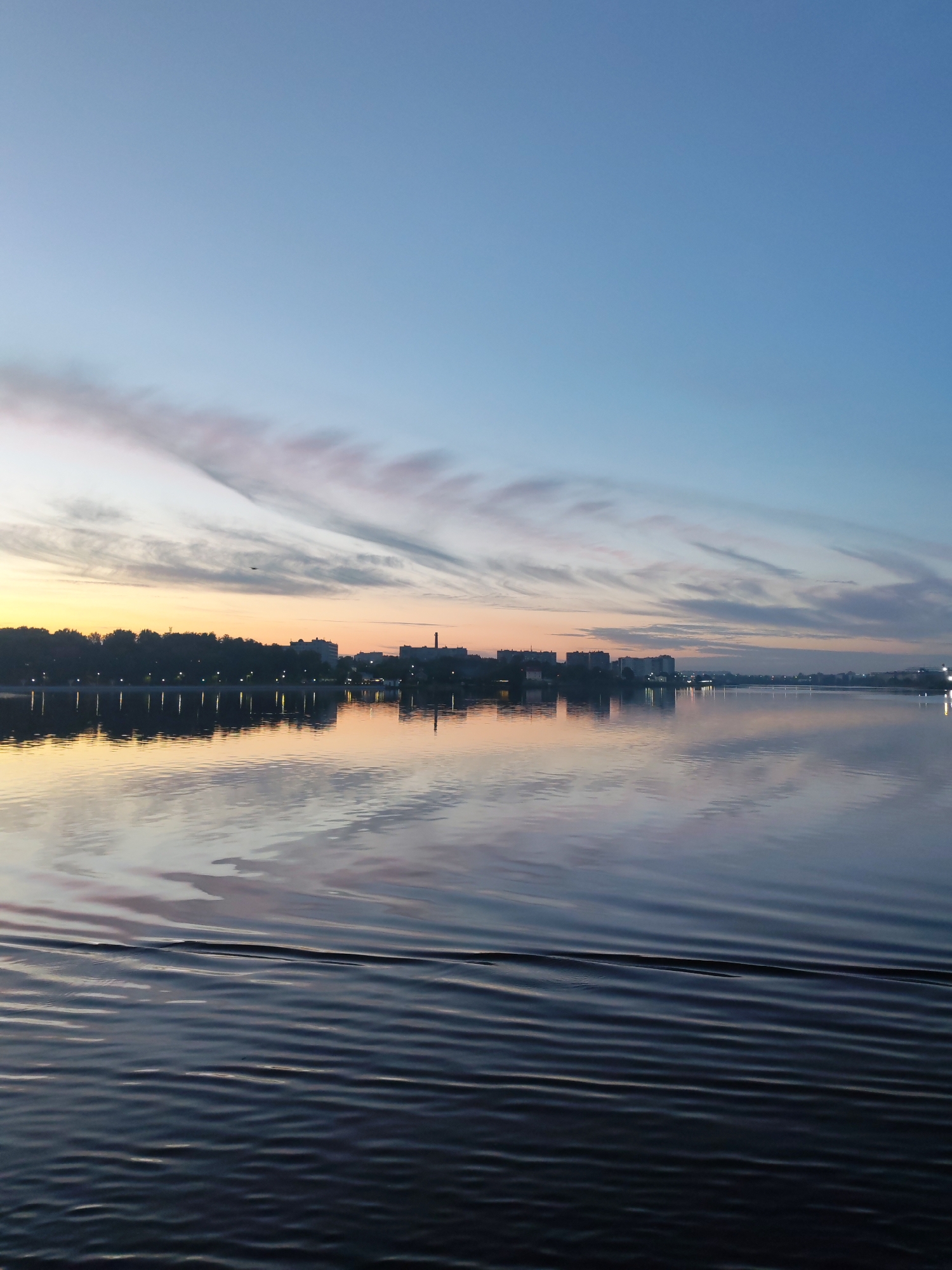 Here's a detailed description of the image:
**Scene:** A serene waterscape at dusk or dawn, likely in Cherepovets, Russia, as suggested by the context. The focus is on a large body of water (likely the Sheksna River or Rybinsk Reservoir), with calm ripples disturbing the surface. The opposite shore presents a cityscape silhouette against a soft gradient sky.
**Environment:** The sky transitions from a deep blue at the top to orange/yellow at the horizon. Wispy clouds stretch horizontally across the sky, reflecting subtle pink and grey hues. The water mirrors the colors of the sky and the shoreline.
**Objects:** A row of trees lines the shore, interspersed with buildings of varying heights, creating a skyline. Artificial lights illuminate the edge of the shore and are reflected in the water. There appears to be a smokestack.
**Details:** The water surface displays a uniform pattern of ripples. The lighting suggests either sunrise or sunset.
**Overall Impression:** The image evokes a sense of tranquility and natural beauty juxtaposed with the presence of an urban environment.