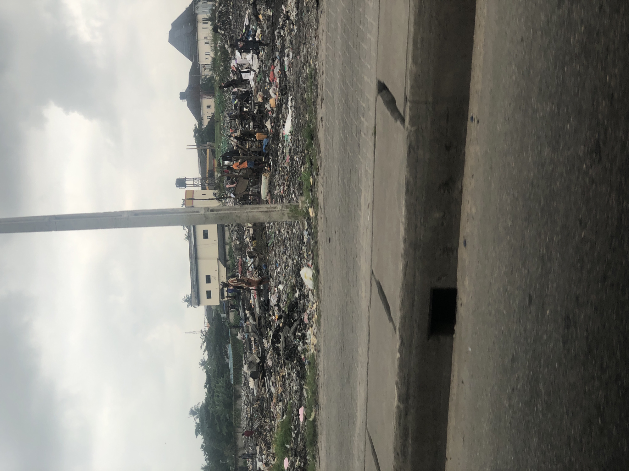 In Satellite Town, Nigeria, a large, open area is filled with debris and refuse, appearing to be a dump site. Numerous individuals are scattered throughout the trash, some appearing to sift through it. In the background, several buildings are visible, including a pale yellow structure with dark windows and a darker building with a peaked roof. There are also trees lining the perimeter of the area. The sky above is overcast and gray. In the foreground, a concrete barrier with visible seams separates the dump site from a road.