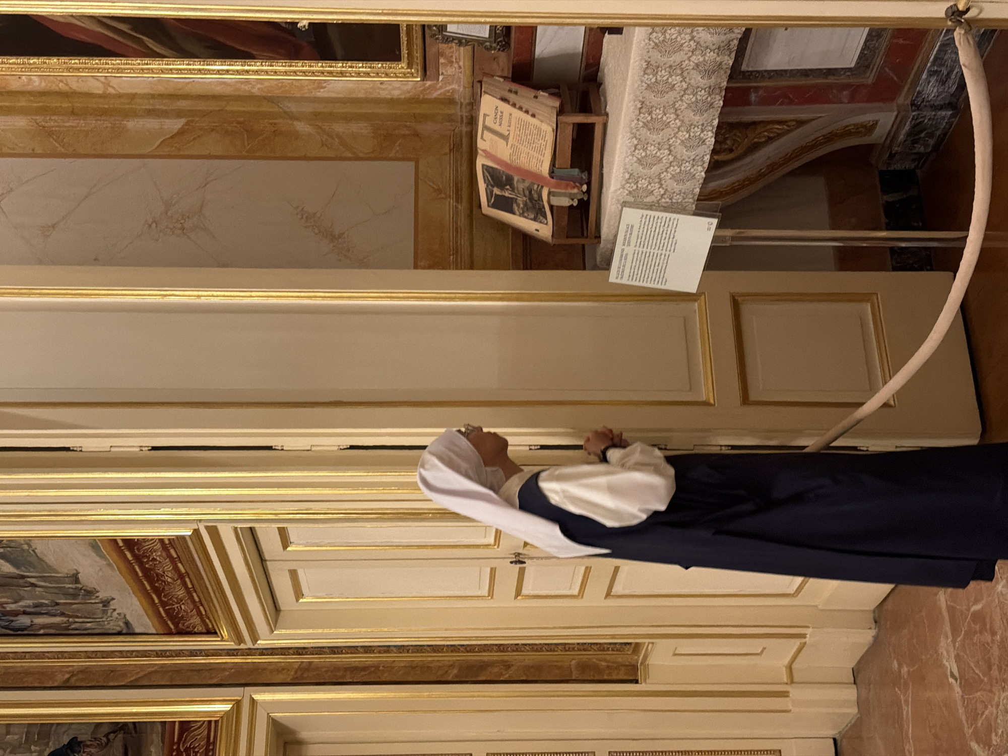 The media file captures an indoor scene within what appears to be a religious or historical setting, possibly in San Lorenzo De El Escorial, Spain, as indicated by the context. A person dressed in a nun's habit, consisting of a white wimple and a dark blue robe, is the central subject. The nun is standing and looking upwards with her hands clasped in front of her chest, suggesting a moment of prayer or contemplation.
Behind the nun, the scene unfolds with elements of ornate architecture. There are richly framed paintings, likely religious in nature, lining the walls. The walls themselves are decorated with elaborate molding and a marbled texture in some areas. In the upper portion of the image, a large, open book rests on a wooden stand. The book appears old and is open to a page with significant text and an illustration. To the right of the book, a white lace fabric is draped, adding to the decorative ambiance. A white informational placard is also visible near the book. The lighting suggests an indoor environment, likely during daytime, with no specific indicators of the weather. The overall atmosphere conveyed is one of solemnity and reverence, consistent with a place of worship or a museum dedicated to religious history.