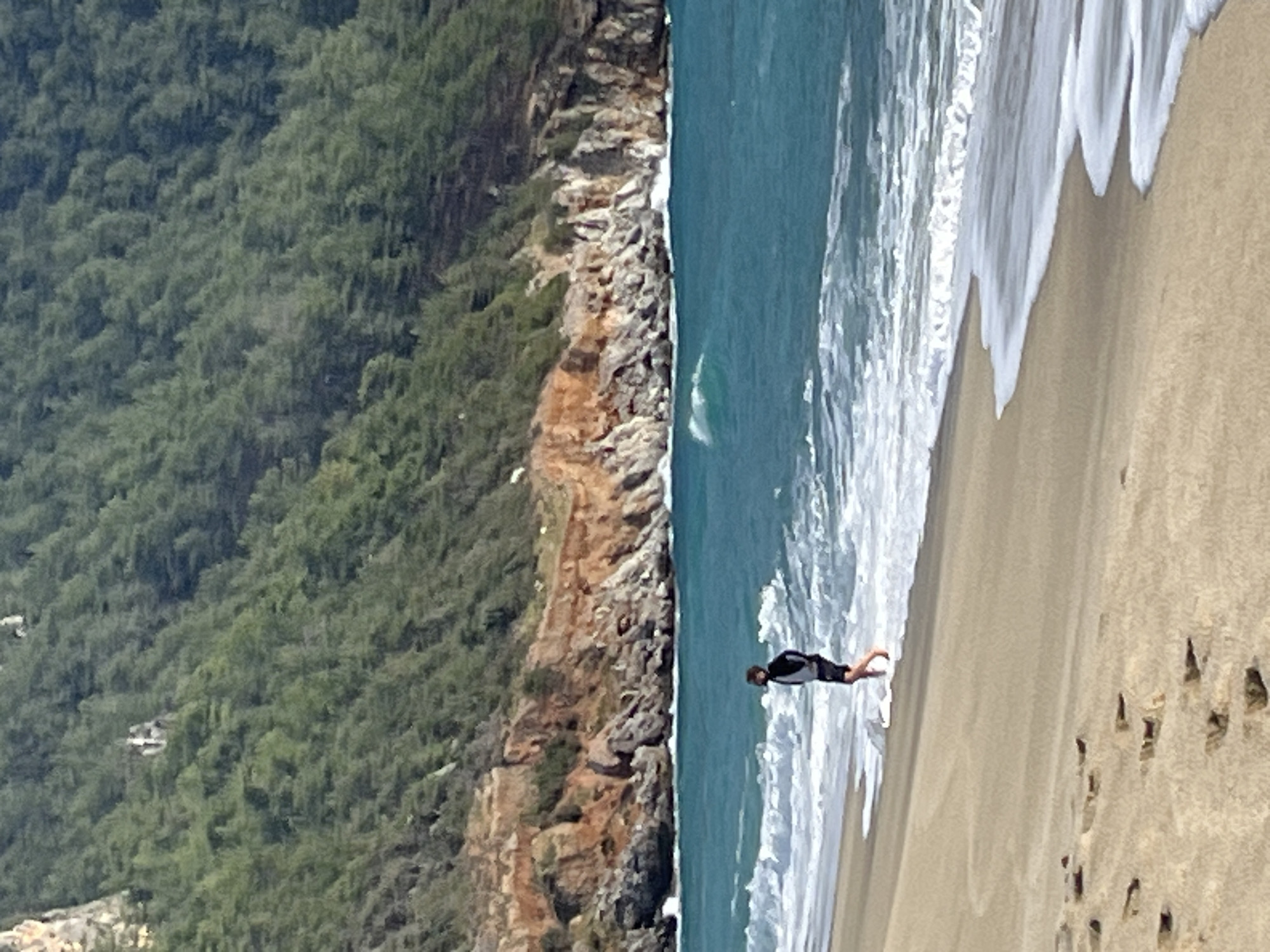 The image depicts a coastal scene at Cleopatra Beach, Alanya, Turkey. In the foreground, a light-colored sandy beach displays multiple footprints. The midground features a lone individual walking barefoot along the waterline, with receding waves around their ankles. This individual wears a dark, possibly two-toned, long-sleeved top and dark shorts, facing slightly towards the right. The ocean is turquoise, with white-capped waves breaking near the shore and further out. The background consists of reddish-orange rocky cliffs rising directly from the sea, transitioning into a steep mountain densely covered with dark green conifer-like trees, which extend upwards to fill the top portion of the frame.