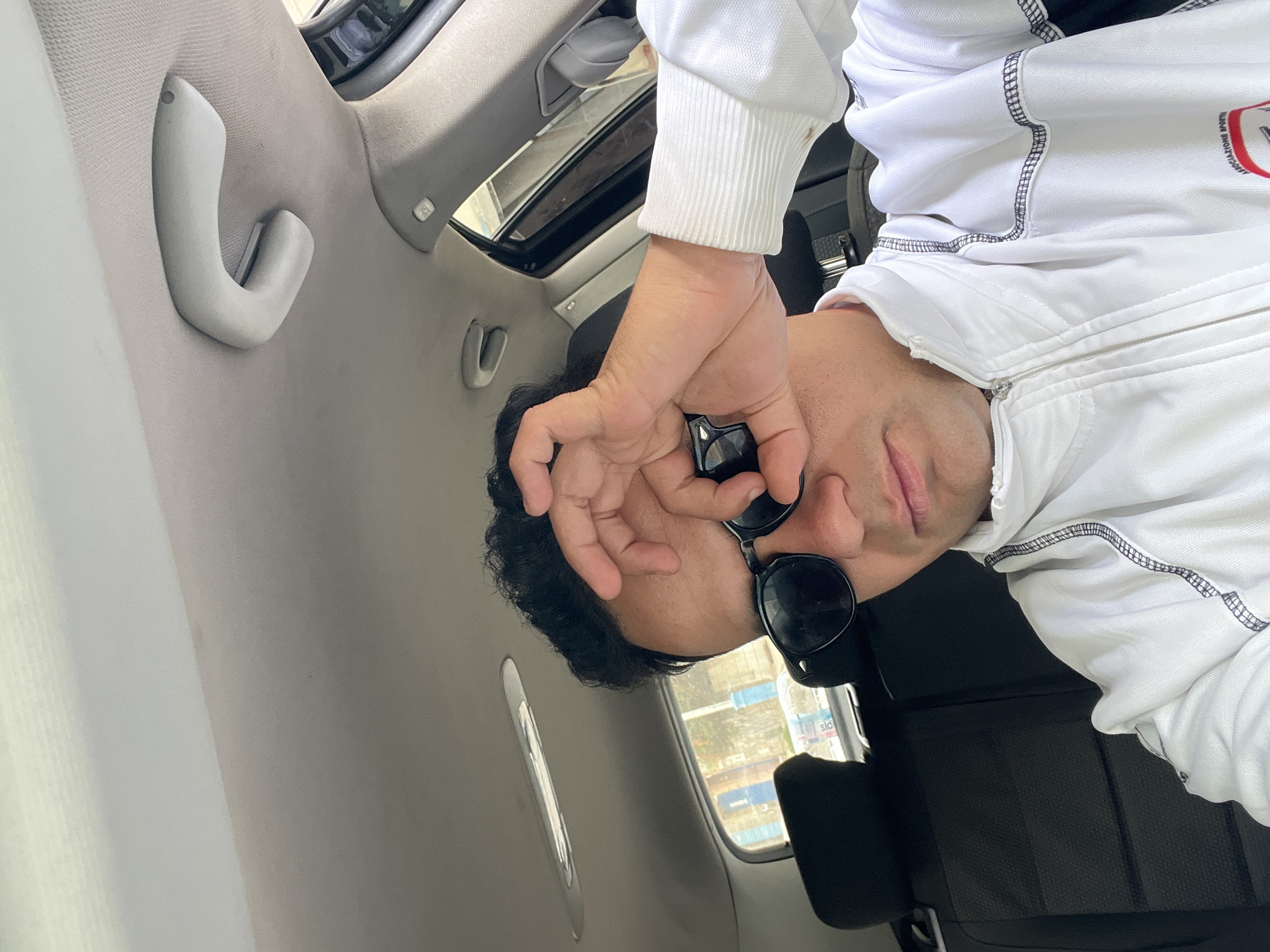 The image, presented rotated 90 degrees clockwise, captures a man inside a car during what appears to be daytime. The scene is likely in Martil, Morocco.
The primary subject is a man, positioned on the right side of the frame, looking towards the viewer. He has short dark hair and light skin. He is wearing dark sunglasses, which he is adjusting or holding with his right hand near his eye. His left hand is not clearly visible. He is dressed in a white long-sleeved jacket or hoodie, featuring black stitching details and a visible zipper. His lips are slightly parted, and he has a neutral expression.
The interior of the car is visible around him, featuring a light grey fabric headliner and side pillars. A light grey plastic grab handle is prominently seen on the ceiling, with another partial handle in the background. A sunroof is visible above, suggesting an open or clear sky. Black car seats are partially visible in the background on the lower left.
Through the car windows, an outdoor environment can be seen. There are faint hints of buildings and possibly greenery. A notable detail outside the window is a blue object, possibly a container, with the partial text "slo" or "sio" visible on it. The bright light streaming into the car suggests a sunny day.
