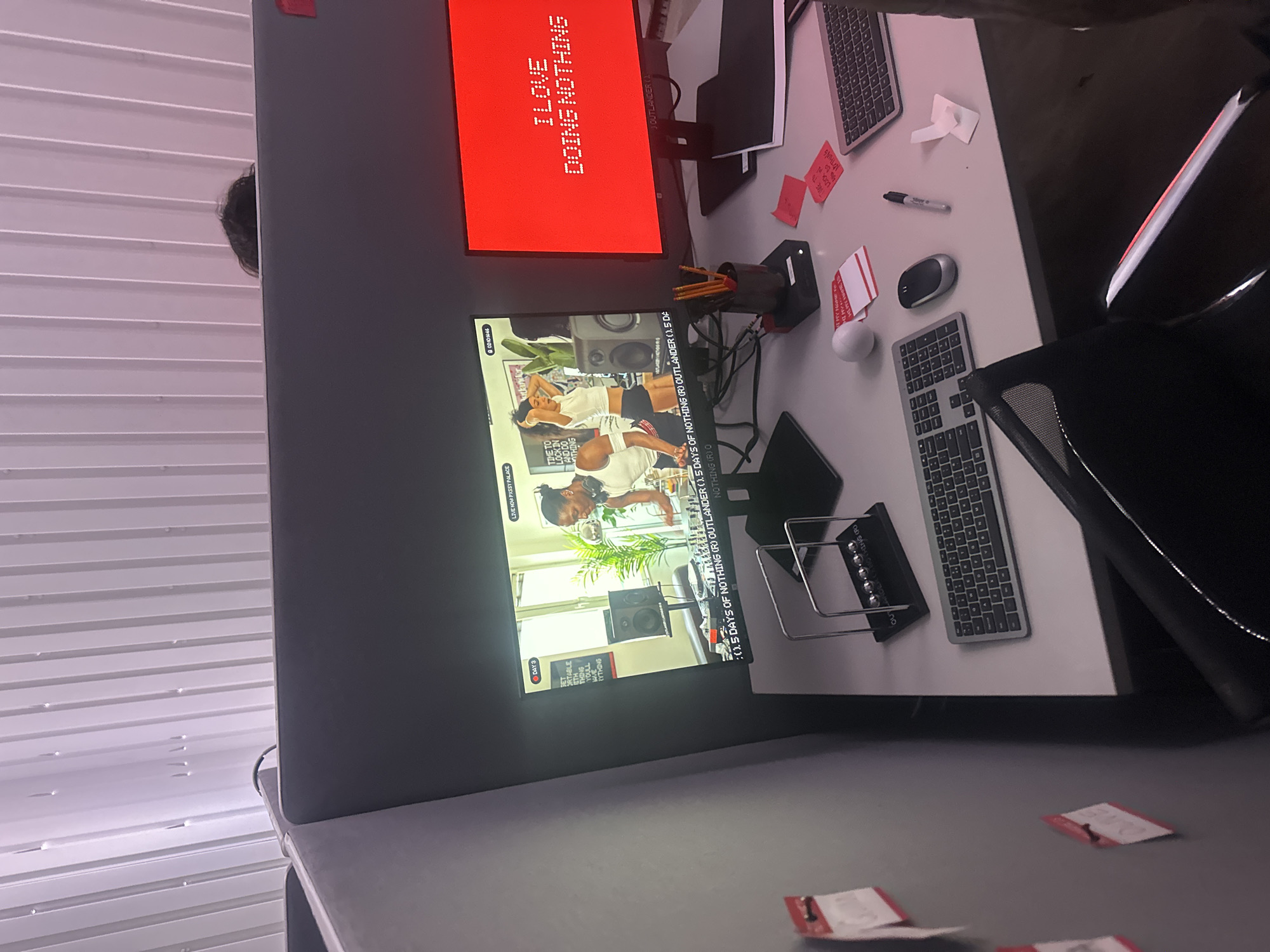 The scene depicts an active office workstation located in London, United Kingdom. A light grey desk is centrally positioned between grey office partitions. On the desk, two monitors are present. The left monitor displays a video featuring two individuals in an interior setting, one female DJ wearing headphones behind a mixer, and a partially visible male. Text overlays include "LIVE NOW PASSY PALACE," "DAY 3," and a scrolling ticker indicating "1.5 DAYS OF NOTHING (R) OUTLANDER." The right monitor displays red background with white pixelated text reading "I LOVE DOING NOTHING."
Clustered on the desk are multiple objects: a black Newton's Cradle, two grey wireless keyboards, a white ergonomic mouse, a black wireless mouse, a black marker pen, a black rectangular device holding several yellow pencils in a pot, a stack of white cards with red borders, and several scattered red sticky notes, some with handwritten text. A black notebook or folder rests near the right monitor. The left partition wall has several red sticky notes affixed with pushpins. Behind the workstation, the top of a person's head with dark, curly hair is visible above the partition. The ceiling above features white corrugated metal panels with embedded lights. A black office chair is partially visible in the lower foreground.