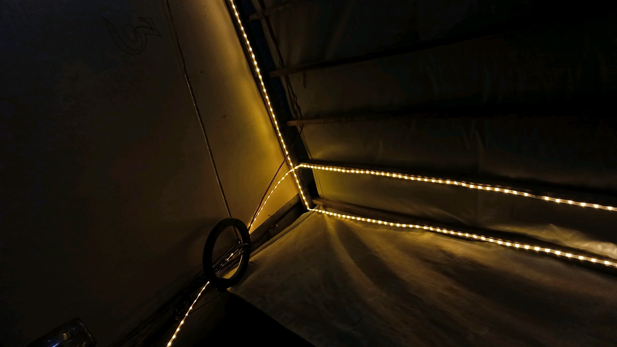 The image captures the interior of what appears to be a tent or a covered structure, illuminated by a string of yellow LED lights. The lights are strategically placed along the wooden beams and fabric walls of the structure, creating a warm and ambient glow. The fabric walls are a light, possibly beige or off-white color, and show creases and folds, indicating they are made of a pliable material. Some of the fabric appears to be a darker color, suggesting different sections or layers within the structure. A circular object, possibly a ring or part of a fixture, is visible on the left side of the image, attached to one of the light strings. There is also a faint, possibly carved or drawn symbol on the upper left section of the fabric. The overall impression is one of a dimly lit, enclosed space with a focus on the decorative lighting. The scene does not provide any specific indicators to pinpoint its location within the city of Adi Luhur, Indonesia, or any details about people, actions, or interactions.
