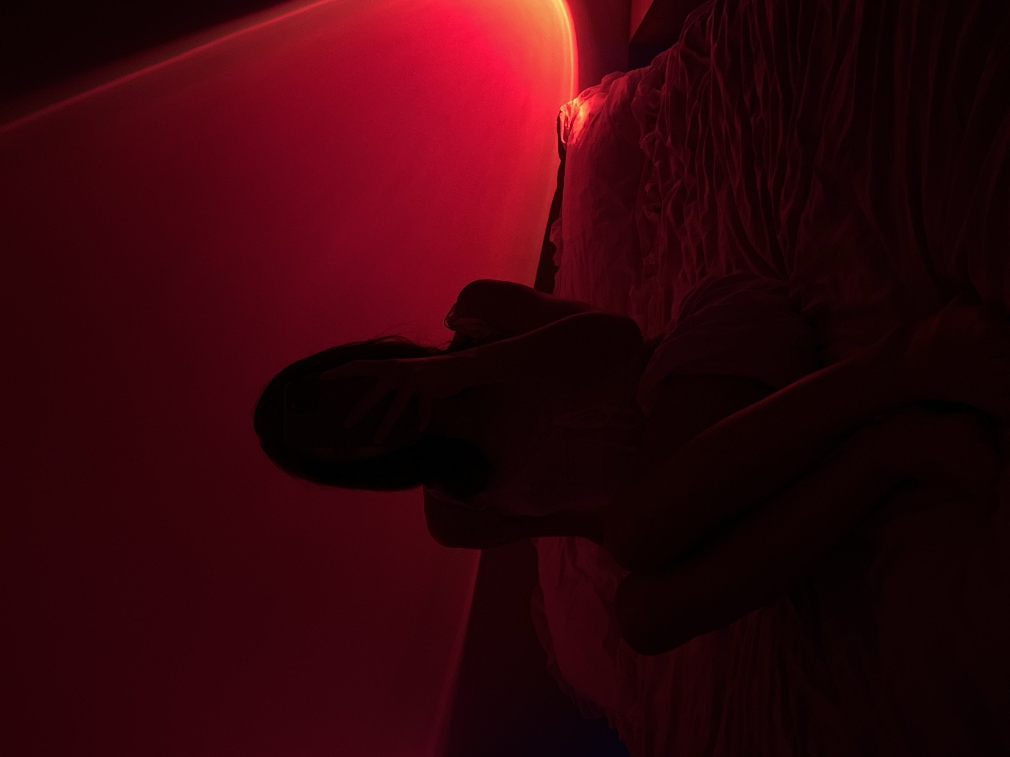 In a dimly lit room, a person is seated on a bed, their face obscured by a phone they are holding. The environment is bathed in a vibrant red light, likely from a sunset lamp, casting a warm glow on the surroundings. The bed appears to be covered in rumpled sheets, suggesting a relaxed or intimate setting. The person is wearing what seems to be a light-colored top. The image does not provide any specific information about the location within the city of Ternopil, Ukraine.