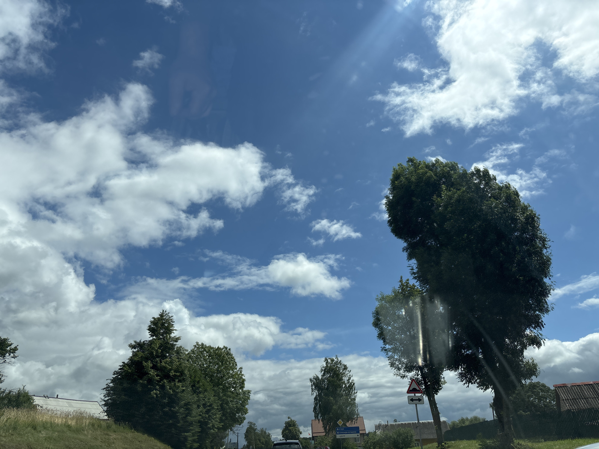 The image depicts a rural road scene in the vicinity of Капыльскі сельскі Савет, Belarus. The sky is bright blue with scattered white cumulus clouds and visible sunbeams. Several large, mature trees with dense green foliage line the road. In the foreground, to the left, a grassy embankment slopes upwards, with a white-roofed building partially visible behind it. Further down the road, on the right side, a dark fence runs alongside the properties, with houses and more trees visible beyond. Traffic signs are present, including one indicating a speed bump with a double-headed arrow. A small section of a black car is visible at the bottom center of the frame, suggesting movement or stationary presence on the road. The overall impression is a sunny, possibly warm, day in a rural Belarusian landscape.