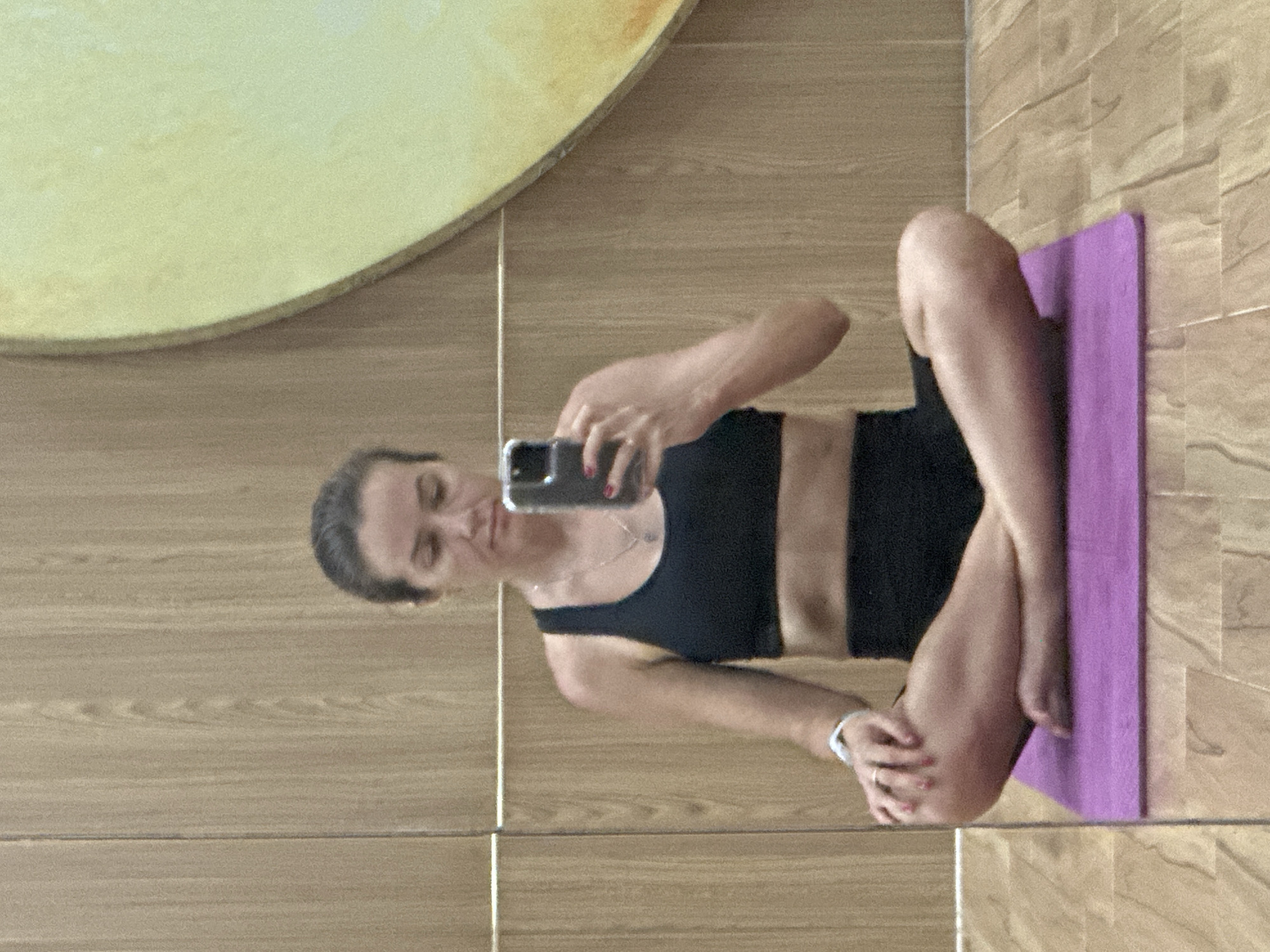 A woman is sitting cross-legged on a purple yoga mat, holding a smartphone to take a mirror selfie. She is wearing a black sports bra and black shorts. Her left arm is bent with her hand resting on her knee, while her right arm is extended to hold the phone. A watch is visible on her left wrist. The background consists of a wooden wall with horizontal lines and a portion of a large, yellowish circular decoration on the right. The floor appears to be made of wood planks. The scene suggests an indoor setting, possibly a gym or a personal workout space, within a room in Bac Nha Trang Ward, Vietnam.