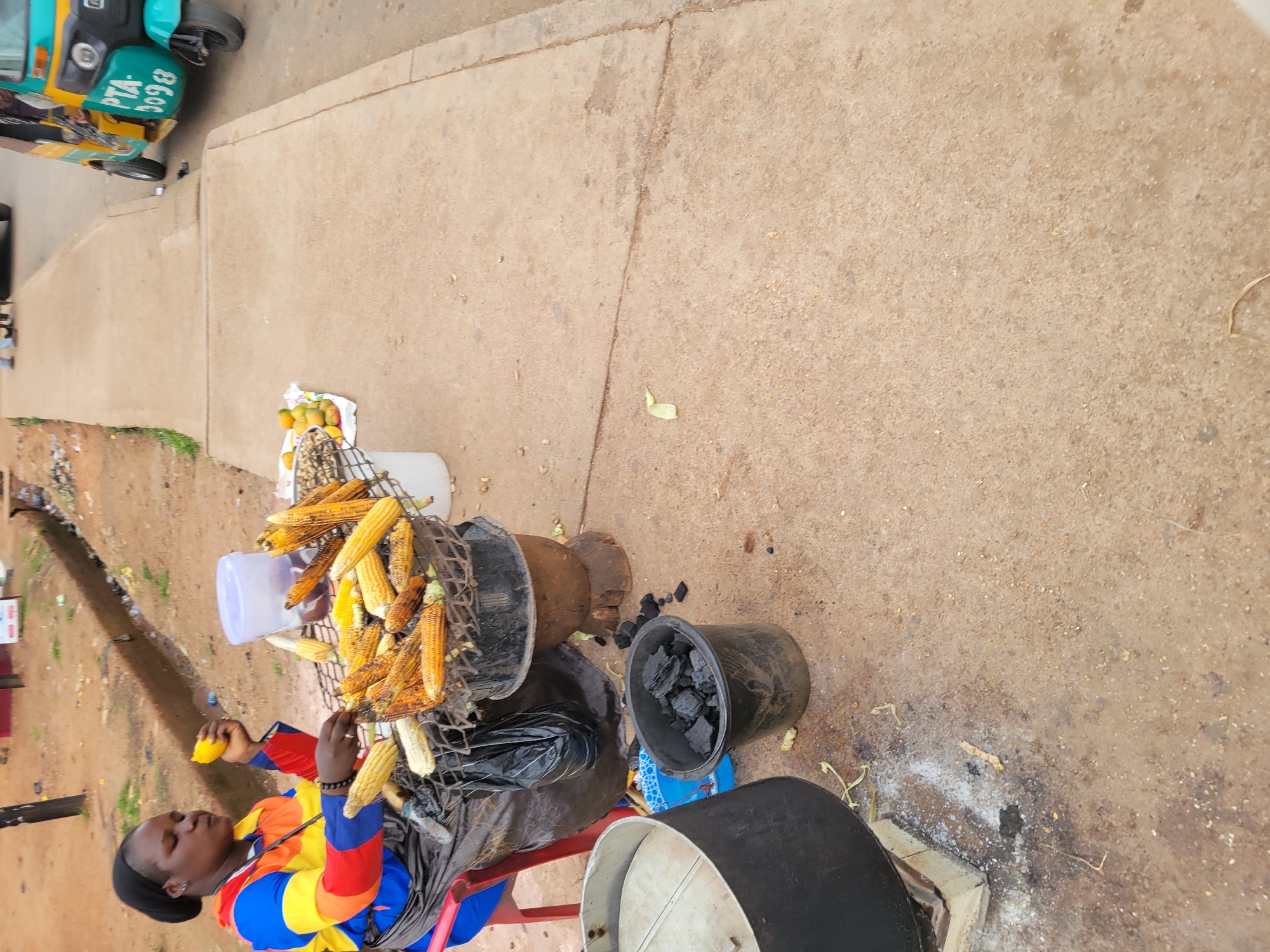 A medium shot shows a woman, viewed from above and to her left, next to a makeshift corn roasting setup along a sidewalk in an unknown city location. The woman has dark skin, dark hair wrapped in a black head covering, and wears a colorful striped shirt with red sleeves. She is holding a piece of fruit, possibly a lemon or orange. Next to her is a metal grate piled with roasted corn on the cob. There's also a white plastic container, a larger pot, a bucket filled with charcoal, and a partially visible metal cooking pot or oven. The ground is dirt mixed with sand. A paved sidewalk extends across the upper portion of the shot, and a blue three-wheeled vehicle is parked on the edge of the sidewalk at the top left. Other buildings are visible in the background.