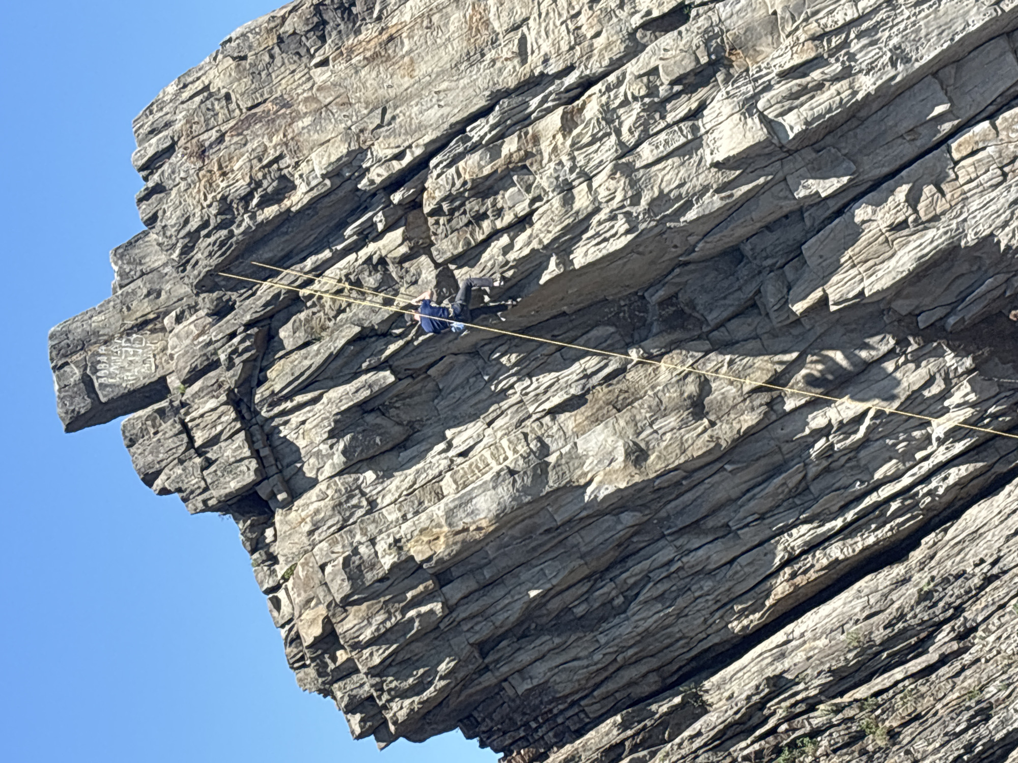 A man in a blue shirt and dark pants is suspended on a taut rope stretched across a rock face. The rock face is composed of light gray, layered rock formations with many cracks and crevices. In the lower right corner, there is some green vegetation growing from the rock. The sky visible behind the rock face is clear and blue. The location is in the city of Vladivostok, Russia.