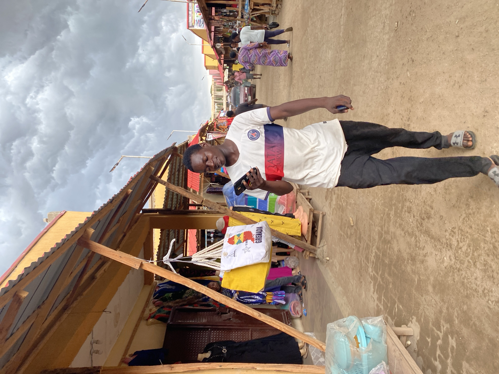 The image depicts a man standing in a marketplace in Kafanchan B, Nigeria. The man is wearing a white t-shirt, black pants, and sandals and is holding a smartphone. He appears to be smiling. The market is characterized by simple, wooden stalls with corrugated metal roofs. Clothing items are displayed for sale. Other people are walking in the background, and a car is also visible. The sky is overcast with heavy, grey clouds. The ground is dusty and appears to be a dirt road or path. A Grenada t-shirt is visible in one of the stalls.