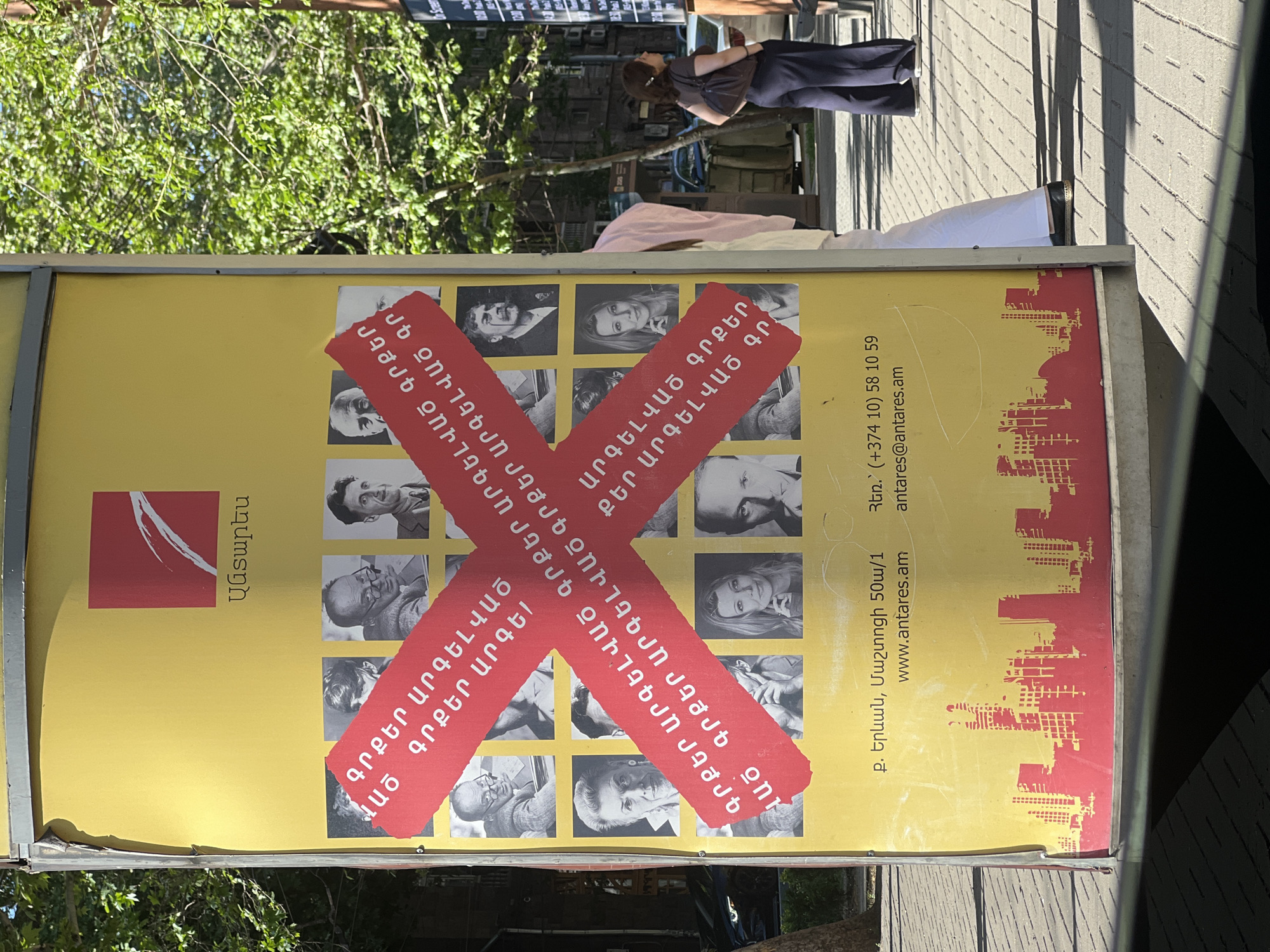 The image shows a vertical outdoor advertisement, likely in Yerevan, Armenia, due to the presence of Armenian text and the mention of the city in the contact information. The advertisement is for "Antares," a brand or organization indicated by its logo and name. The poster has a yellow background with a red cityscape design at the bottom. A red "X" shape is superimposed over a grid of black and white portraits. The text in Armenian is repeated within the red "X". The advertisement lists an address, "p. Yerevan, Mashtotsi 50a/1," a website, "www.antares.am," a phone number, "+374 10 58 10 59," and an email address, "antares@antares.am."
In the background, a woman is standing on the sidewalk, and trees and a building provide environmental context. The scene is likely outdoors during daylight hours.
