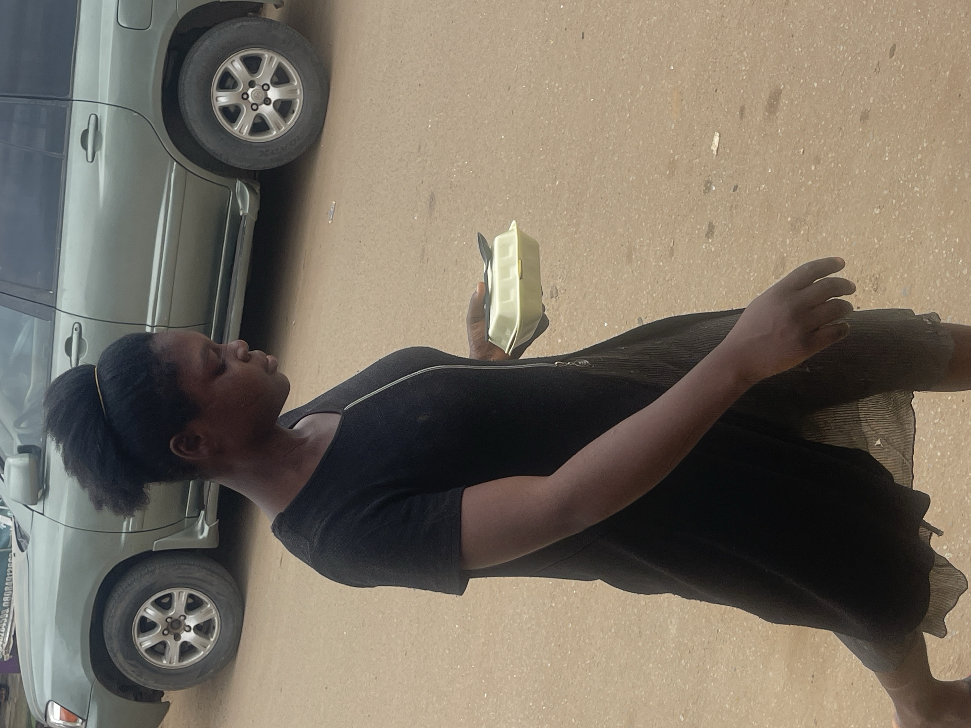 A young woman stands next to a light green SUV, holding a light green rectangular object. She is wearing a black dress with pleats in the skirt and a zipper detail on the upper chest. Her dark hair is pulled back into a ponytail. The SUV is parked on a dusty, paved surface. The scene appears to be outdoors during daylight, likely in Abuja, Nigeria, given the context. There are no clear indicators of the weather, but the light suggests it's not raining. The woman's expression is neutral, and she appears to be looking down at the object she is holding. The object she is holding is a food container, possibly with food inside. The ground is a muted brown color with small speckles of lighter material, consistent with a dirt road or a lightly paved area. The image is oriented vertically, with the woman and car taking up the left and central portions of the frame, and the ground filling the rest.
